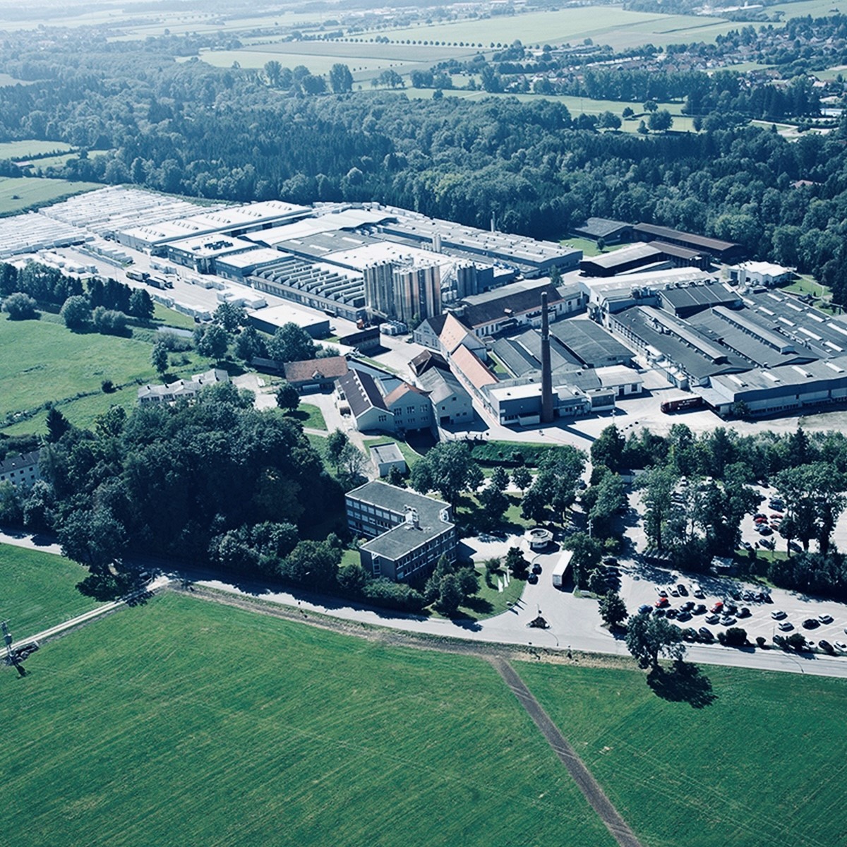 Aerial view of a large industrial complex surrounded by trees and greenery, with a parking lot filled with cars and a wide open field in the foreground.