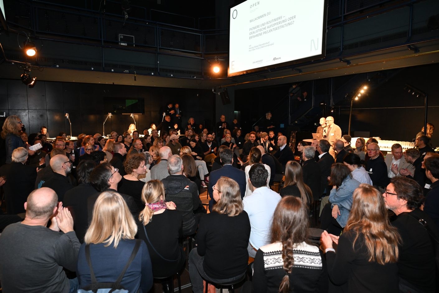 Um grande grupo de pessoas está reunido em uma sala com uma tela grande exibindo texto. Algumas estão sentadas, outras estão em pé e algumas estão aplaudindo. A sala está fracamente iluminada com holofotes no teto e algumas lâmpadas no chão.