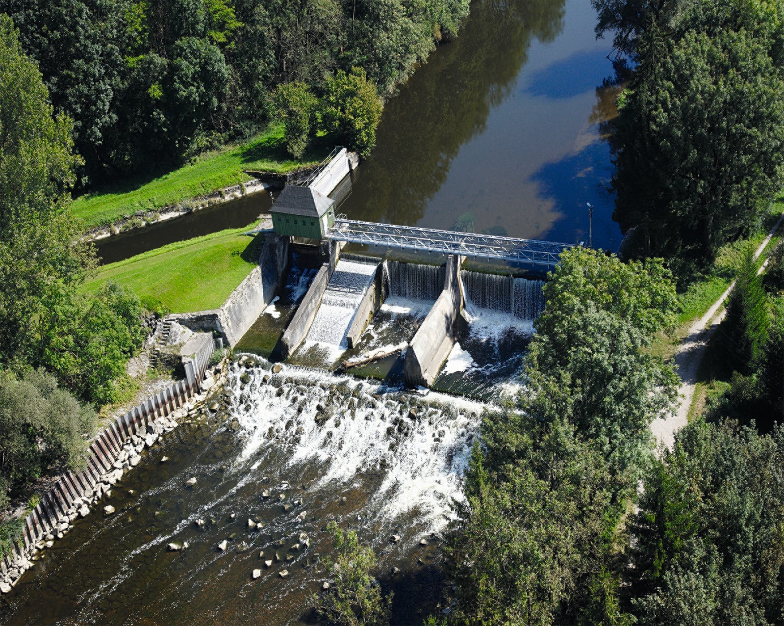 Luftaufnahme eines Damms mit einer Brücke und einem kleinen Gebäude auf der linken Seite, umgeben von Bäumen und Gras, mit einem Fluss, der hindurchfließt.