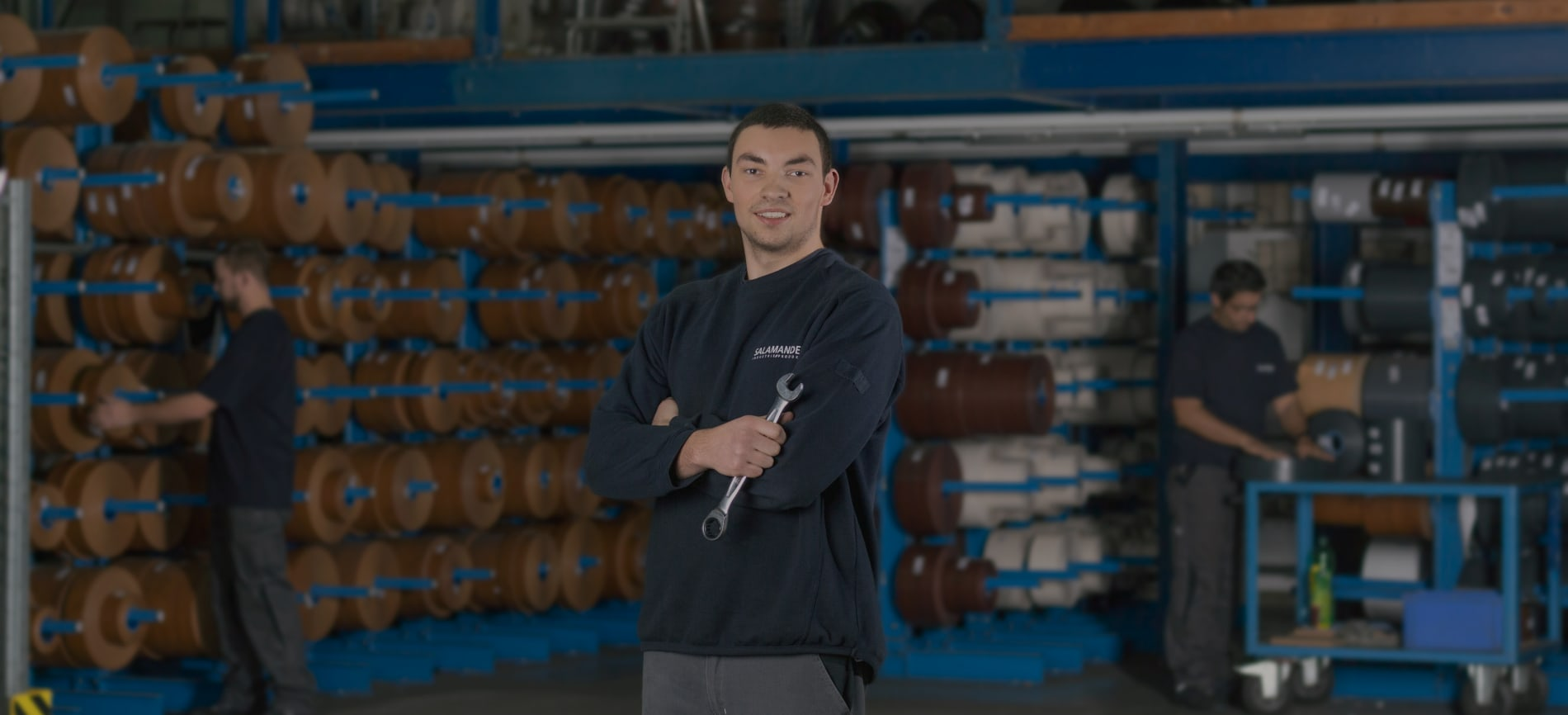 Ein Mann in einem schwarzen Hemd und grauen Hosen steht in einem Lagerhaus mit einem Schraubenschlüssel in den Händen. Hinter ihm arbeiten zwei Männer mit Materialrollen.