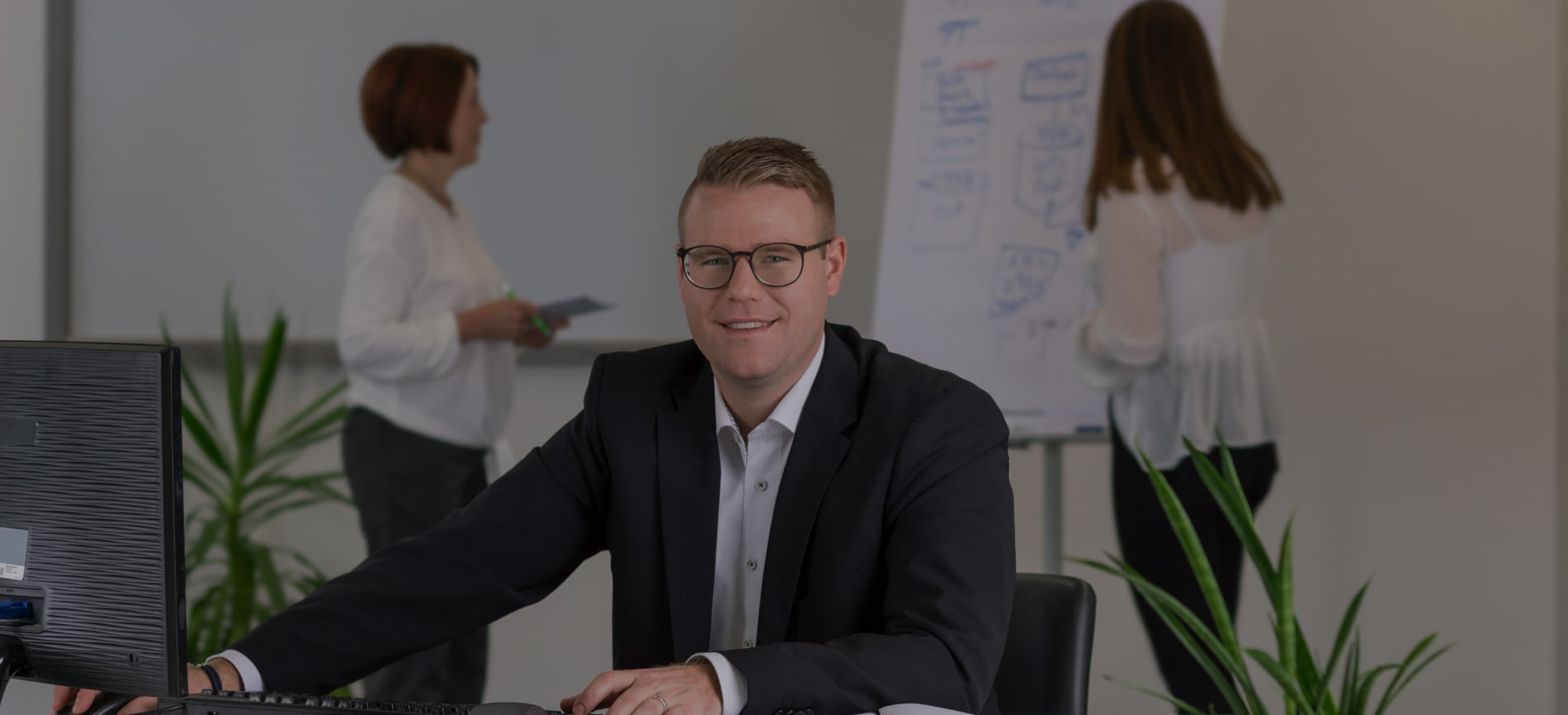 Ein Mann in einem Anzug und mit Brille sitzt an einem Schreibtisch mit einem Computer, während zwei Frauen in weißen Hemden hinter ihm stehen, eine hält einen Stift und Papier.