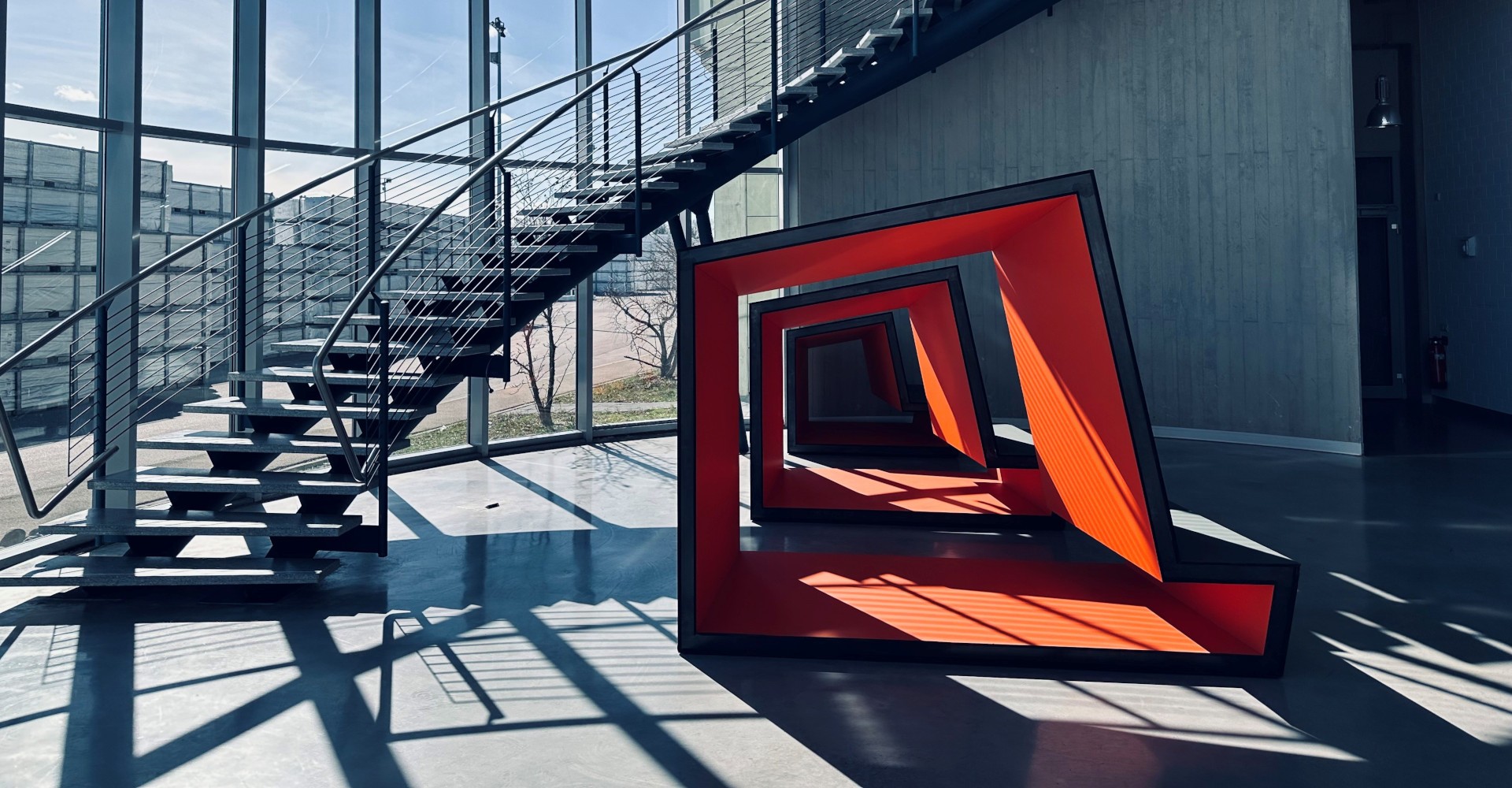A modern building with a spiral staircase and a red geometric sculpture on the floor. The sculpture is made of multiple layers of red and black material, creating a unique and eye-catching design. The building has glass walls and a metal railing, allowing natural light to flood the space. The shadows of the sculpture and staircase are cast on the floor, adding depth and dimension to the scene.