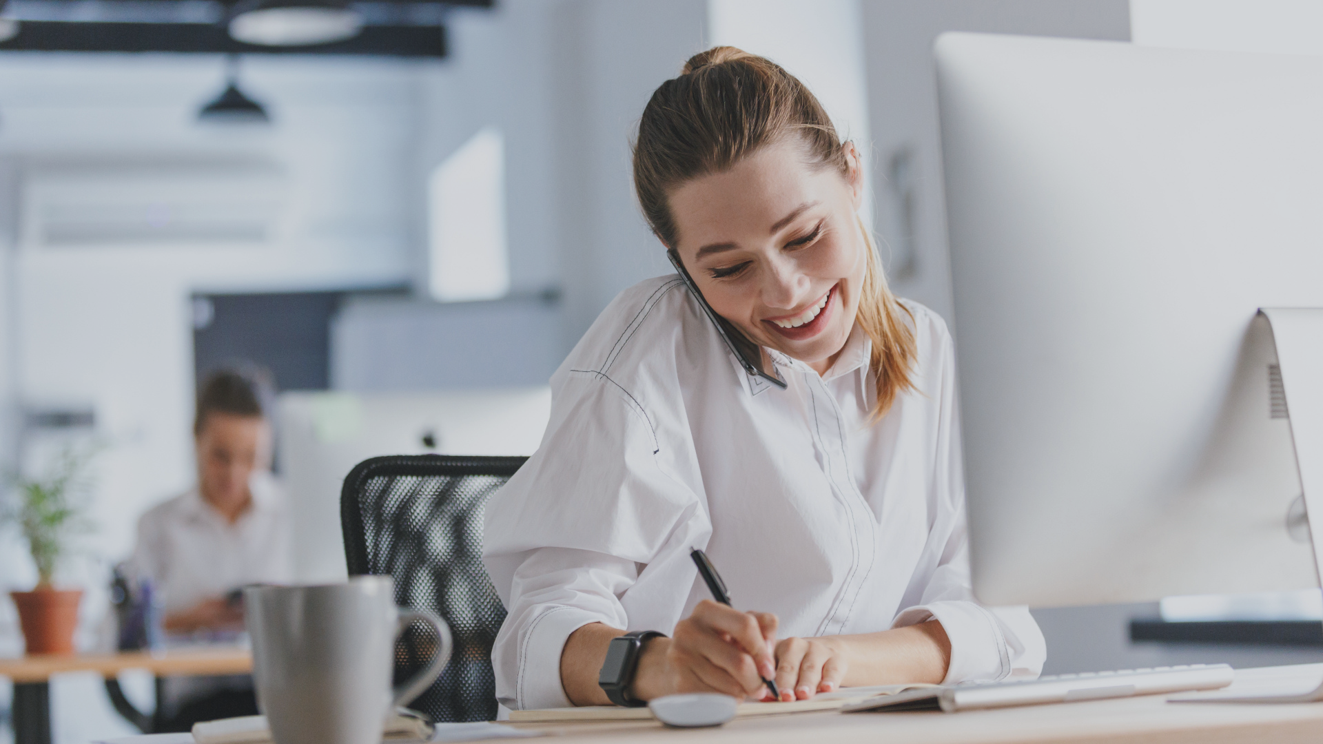 A woman in a white shirt is sitting at a desk in an office, talking on the phone and writing on a piece of paper.