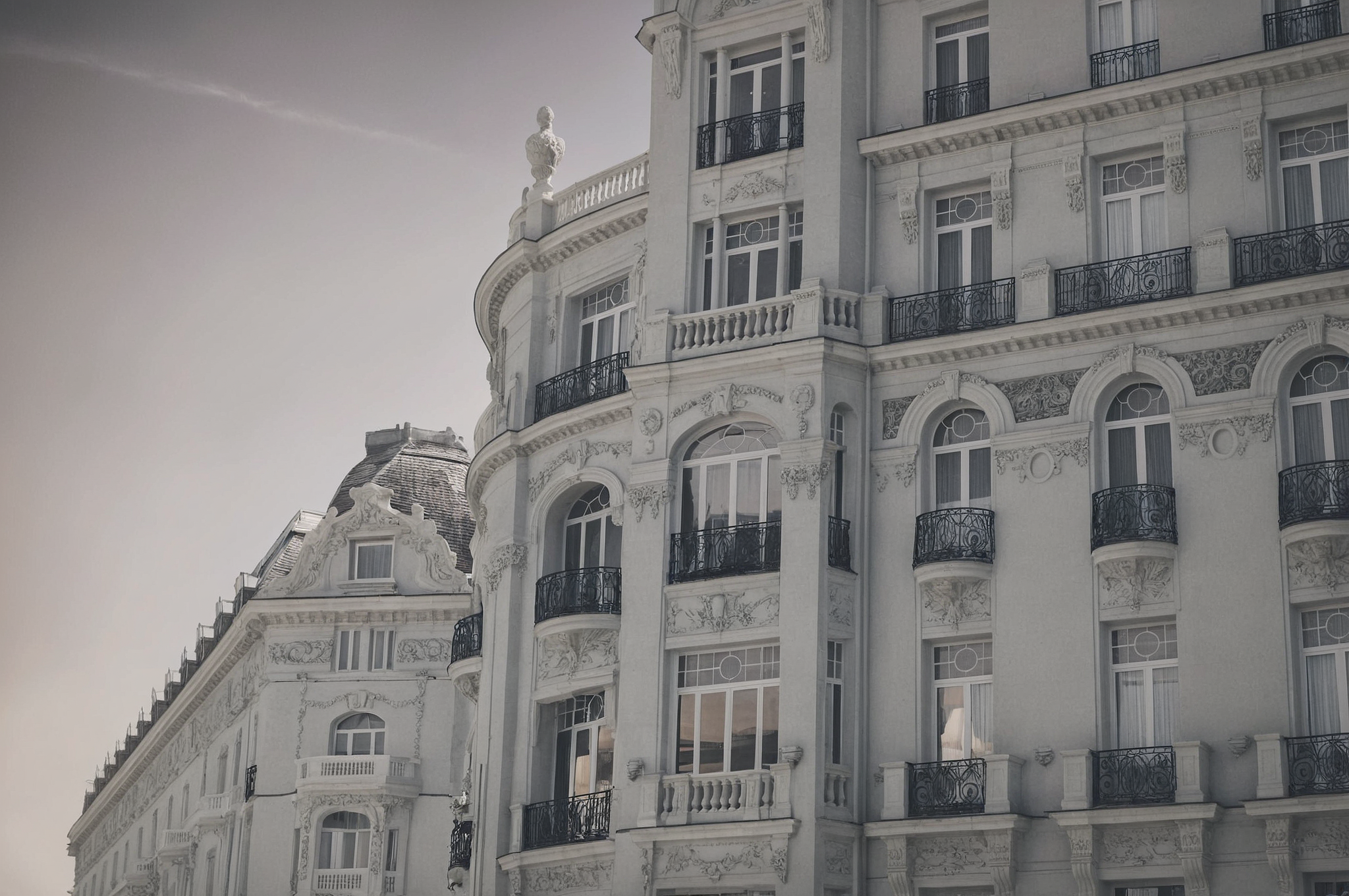 Ein Schwarz-Weiß-Bild eines Gebäudes mit vielen Fenstern und Balkonen. Das Gebäude hat ein geschwungenes Design und ist weiß gestrichen. Die Balkone haben schwarze Geländer und einige von ihnen haben Vorhänge. Das Gebäude hat eine Statue auf seiner Spitze. Das Gebäude ist von anderen Gebäuden umgeben.