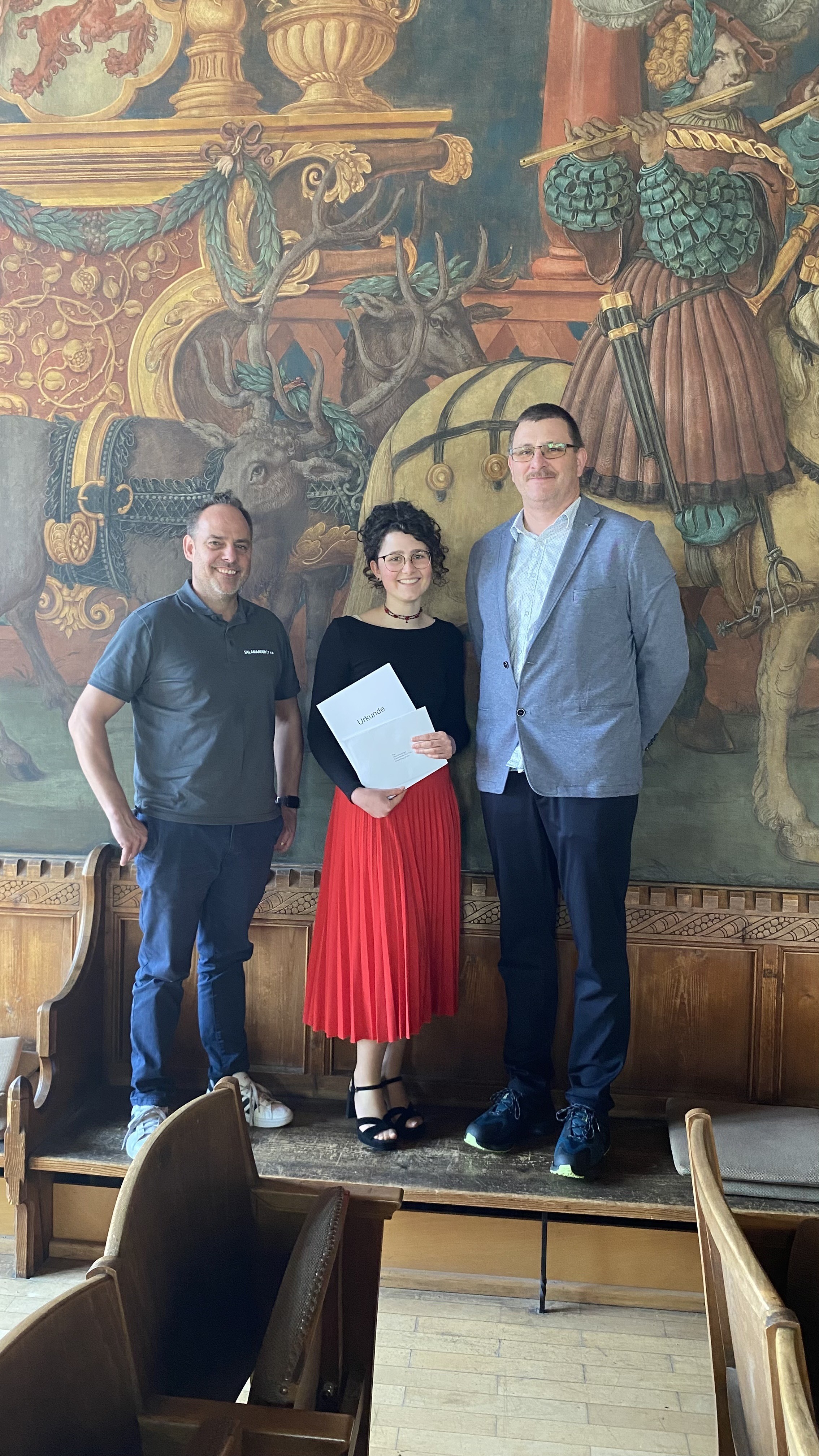Drei Personen stehen vor einem Wandgemälde, eine hält ein Zertifikat, in einem Raum mit Holzstühlen und einer Holzbank.