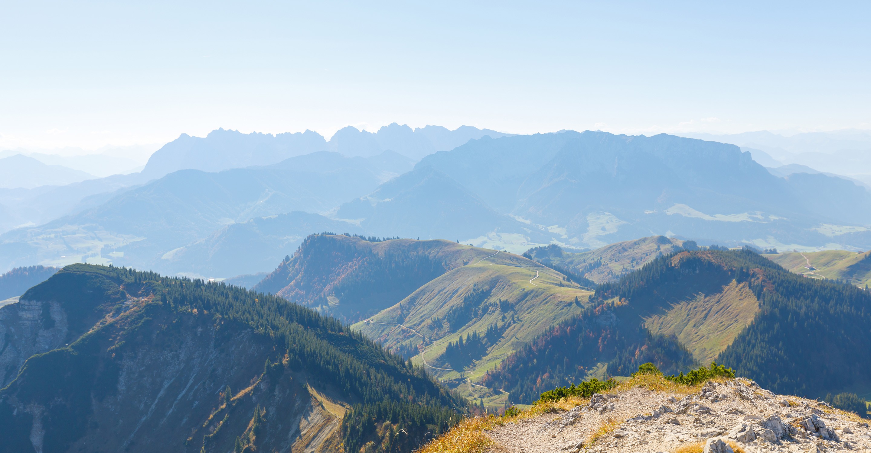 Panoramic view of mountain ranges with lush forests and winding paths under a clear blue sky.