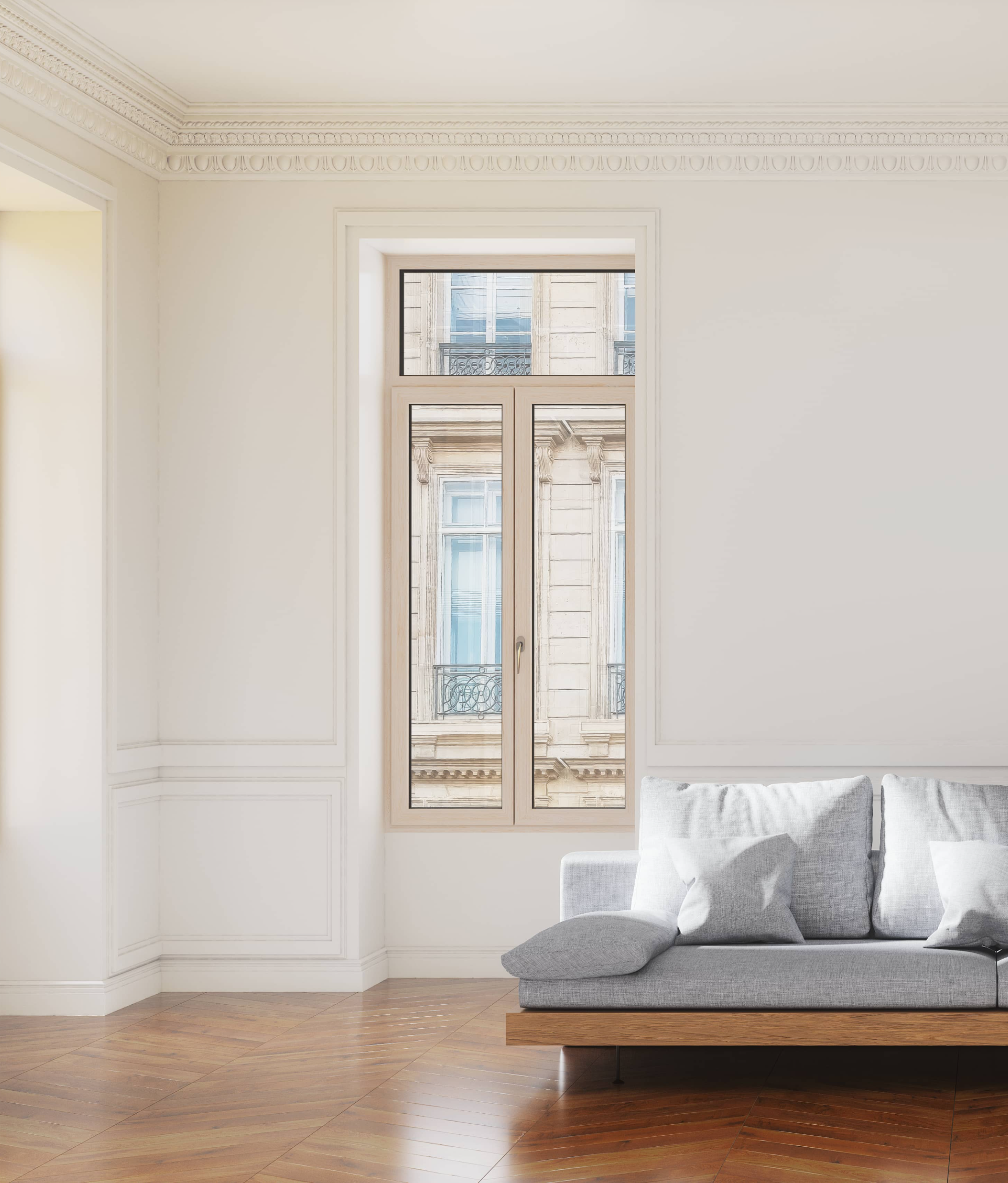 Een grijze bank met witte kussens in een kamer met een raam dat een gebouw met balkons toont.