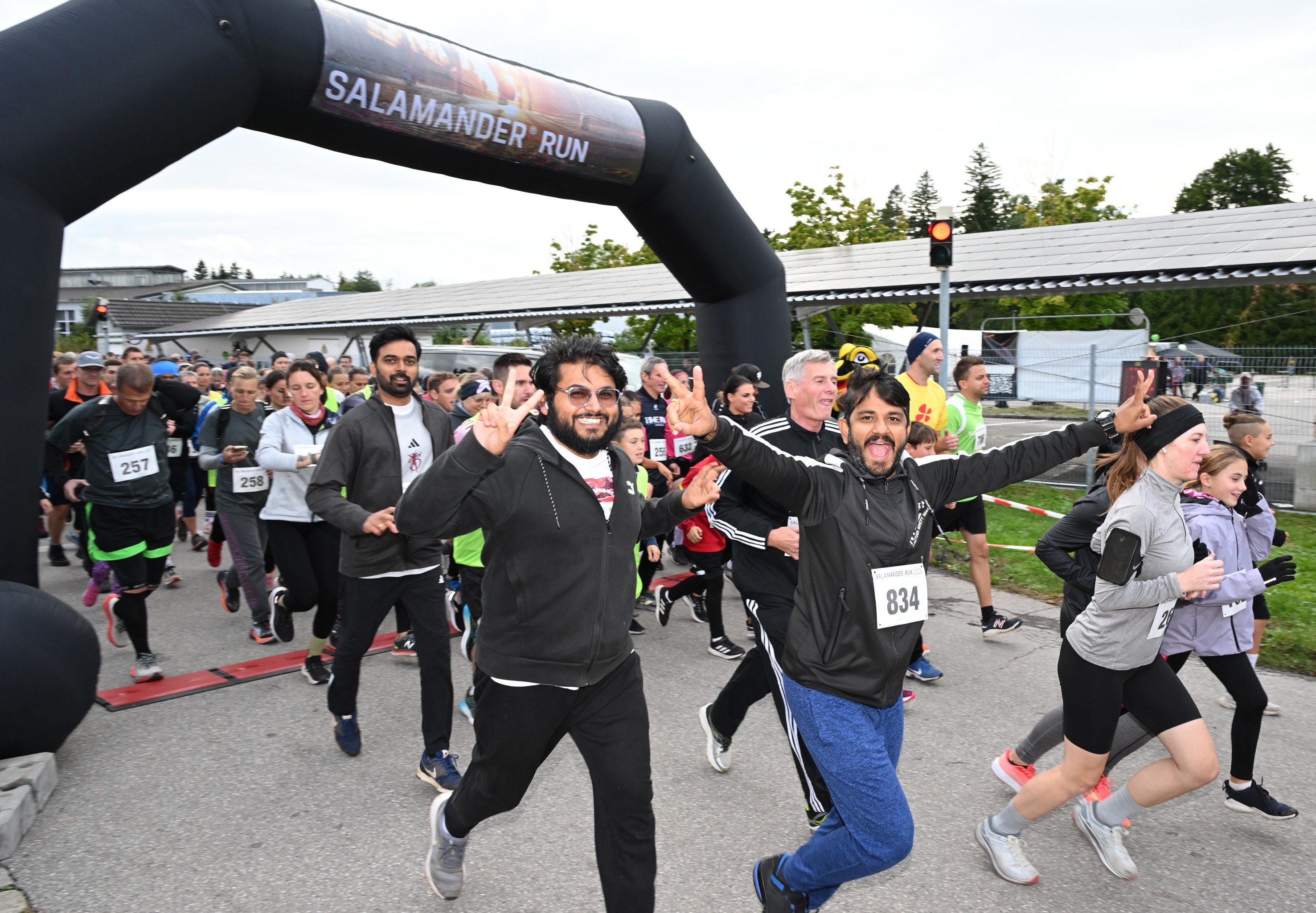 A group of people are running in a marathon. Two men are in the front, wearing black jackets and smiling. They are raising their hands in victory. Behind them are other runners, some of whom are wearing sneakers. In the distance, there is a black inflatable arch with the words "Salamander Run" on it. There are also trees and a building in the distance.