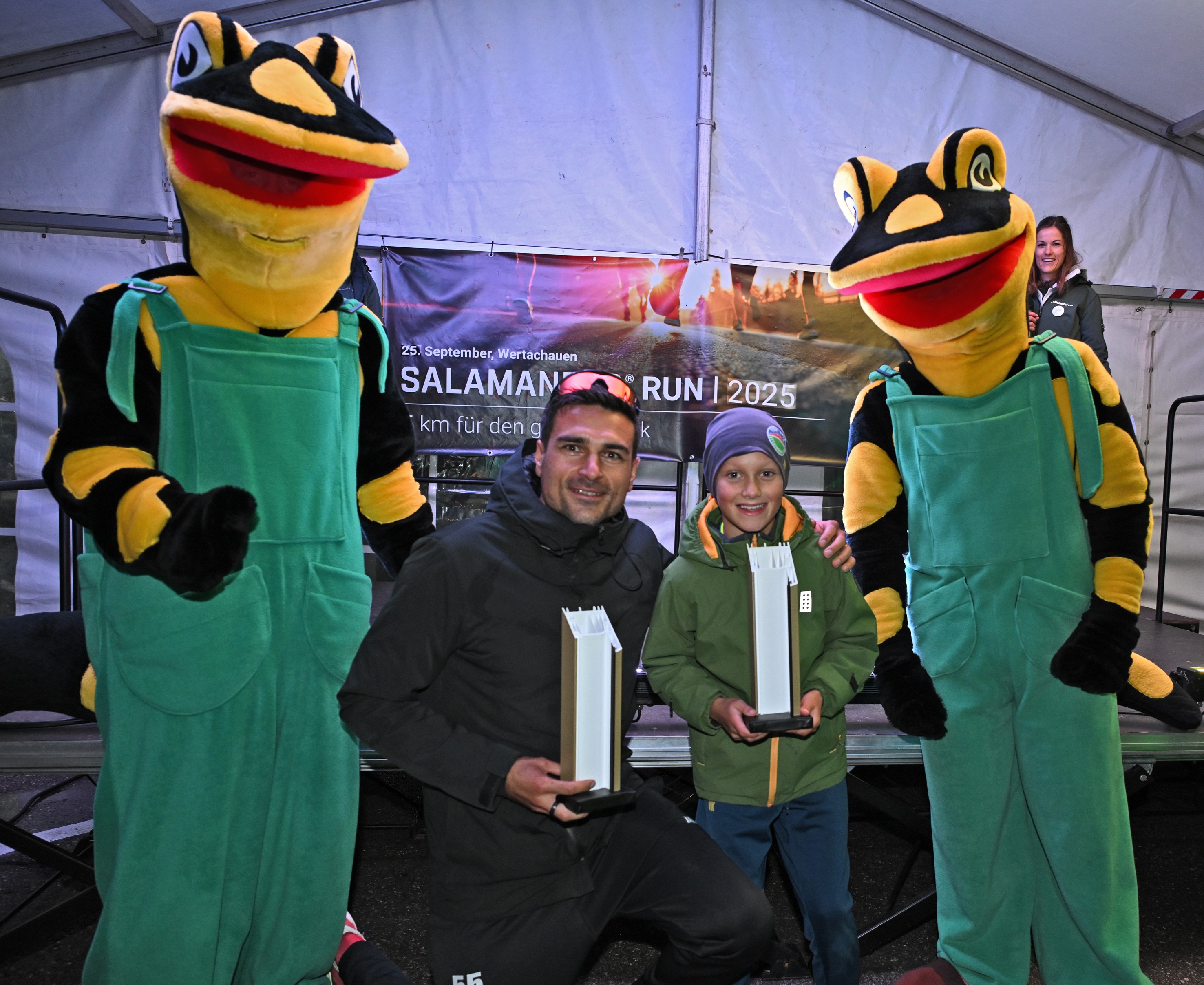 Two mascots, a man, and a boy are posing for a photo. The man and boy are holding trophies.