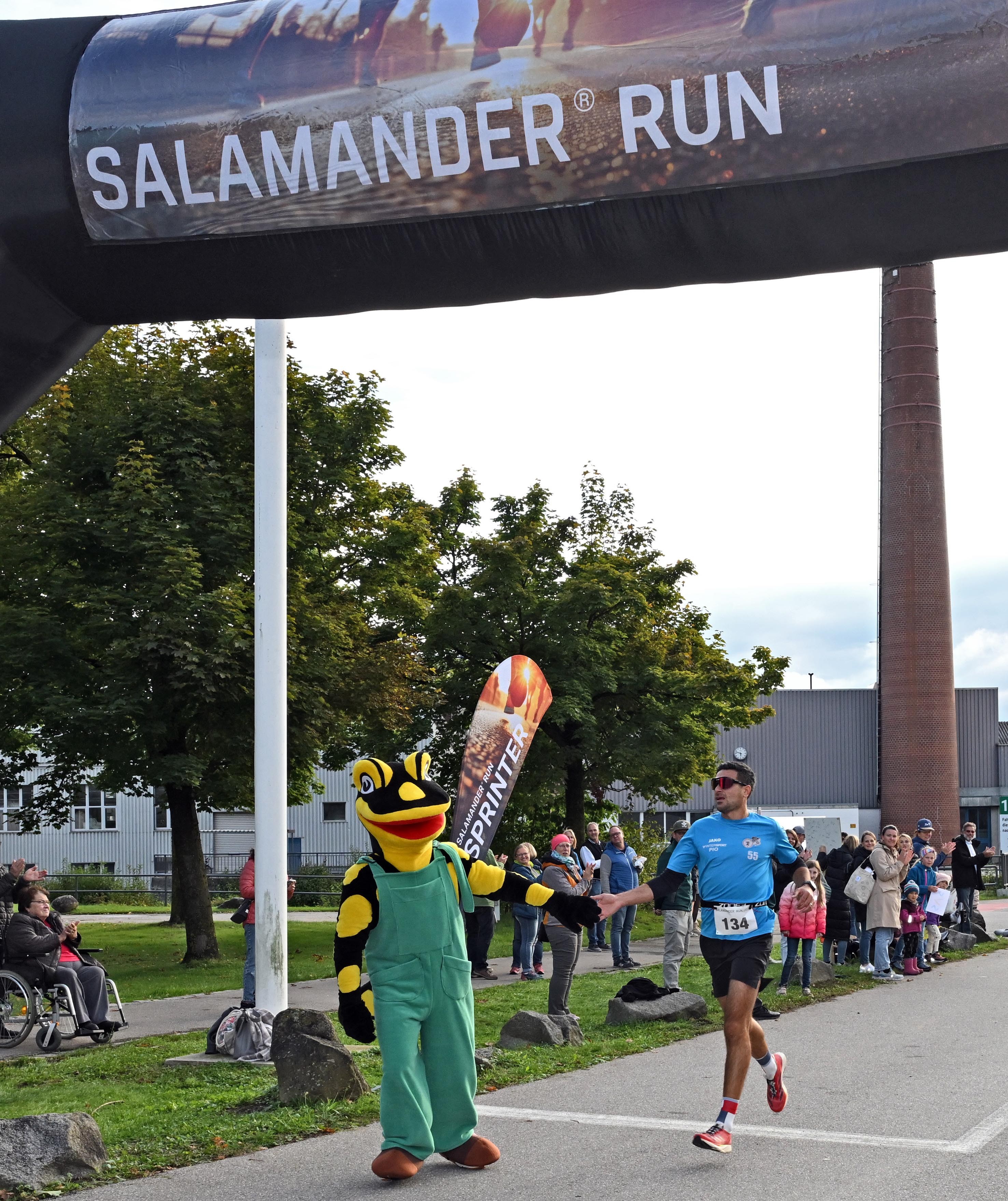 A runner wearing a blue shirt with the number 134 on it is greeted by a frog mascot at the finish line of the Salamander Run.
