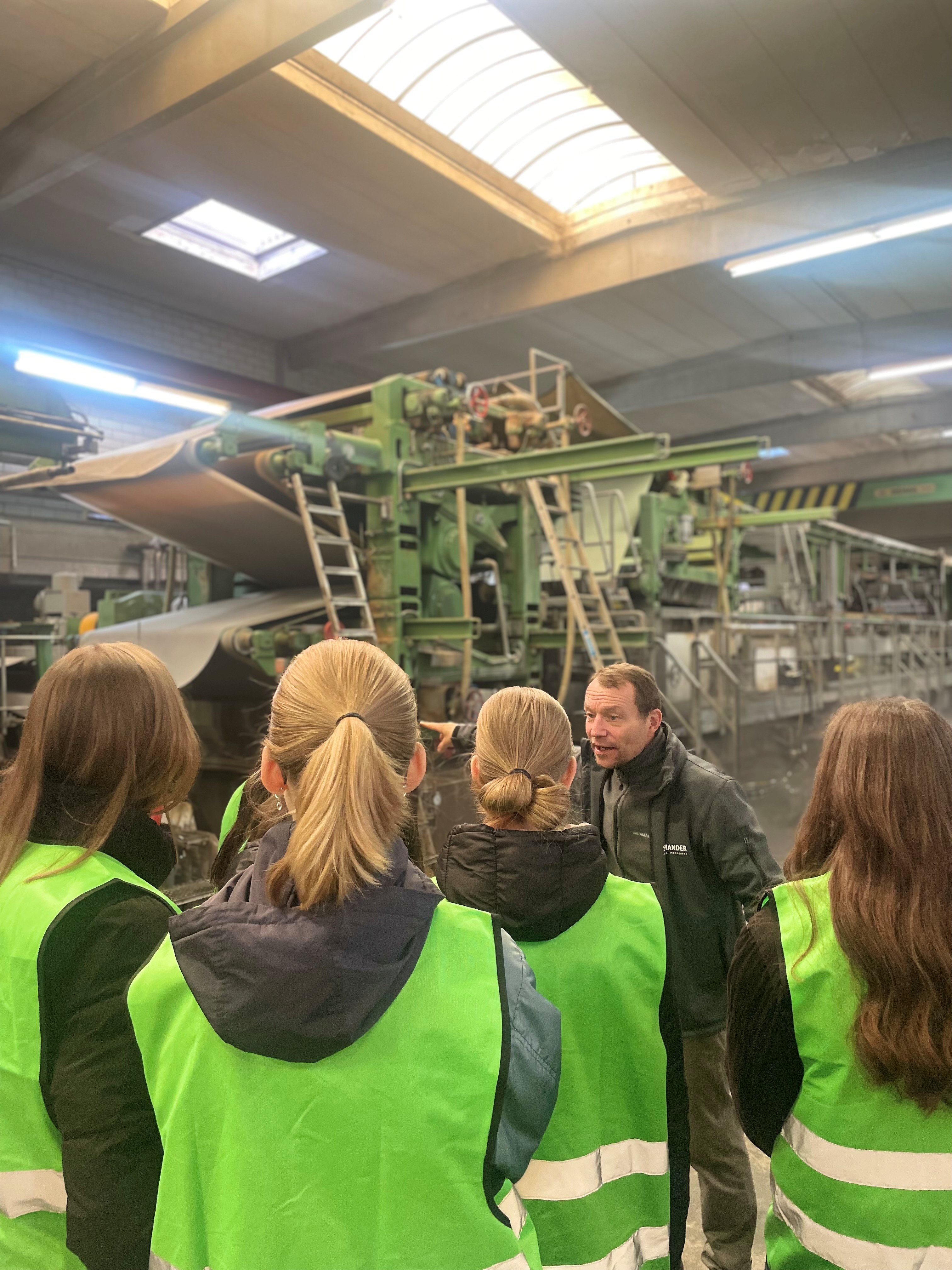 Eine Gruppe von Menschen, die grüne Westen tragen, hat sich um einen Mann in einer schwarzen Jacke versammelt, der auf eine große Maschine in einer Fabrik zeigt.