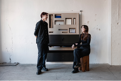 Two people standing and sitting in front of a large piece of furniture in a room.