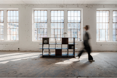 Eine Person, die in einem Raum mit großen Fenstern, einem Regal mit einer Lampe und einem Betonboden geht.