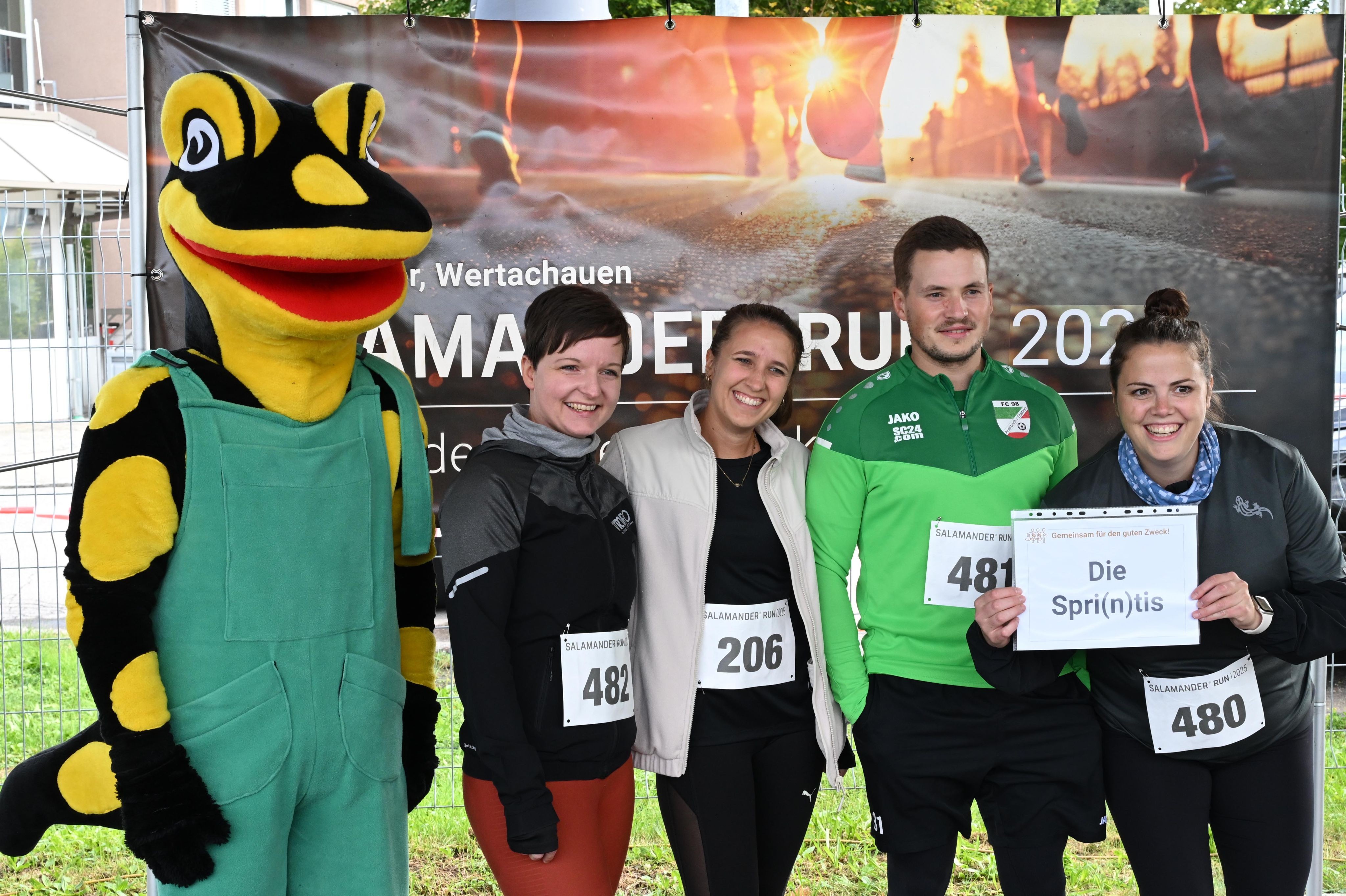 Five people standing together in front of a banner, three of them wearing bibs with numbers, one holding a sign that says "Die Spri(n)tis".