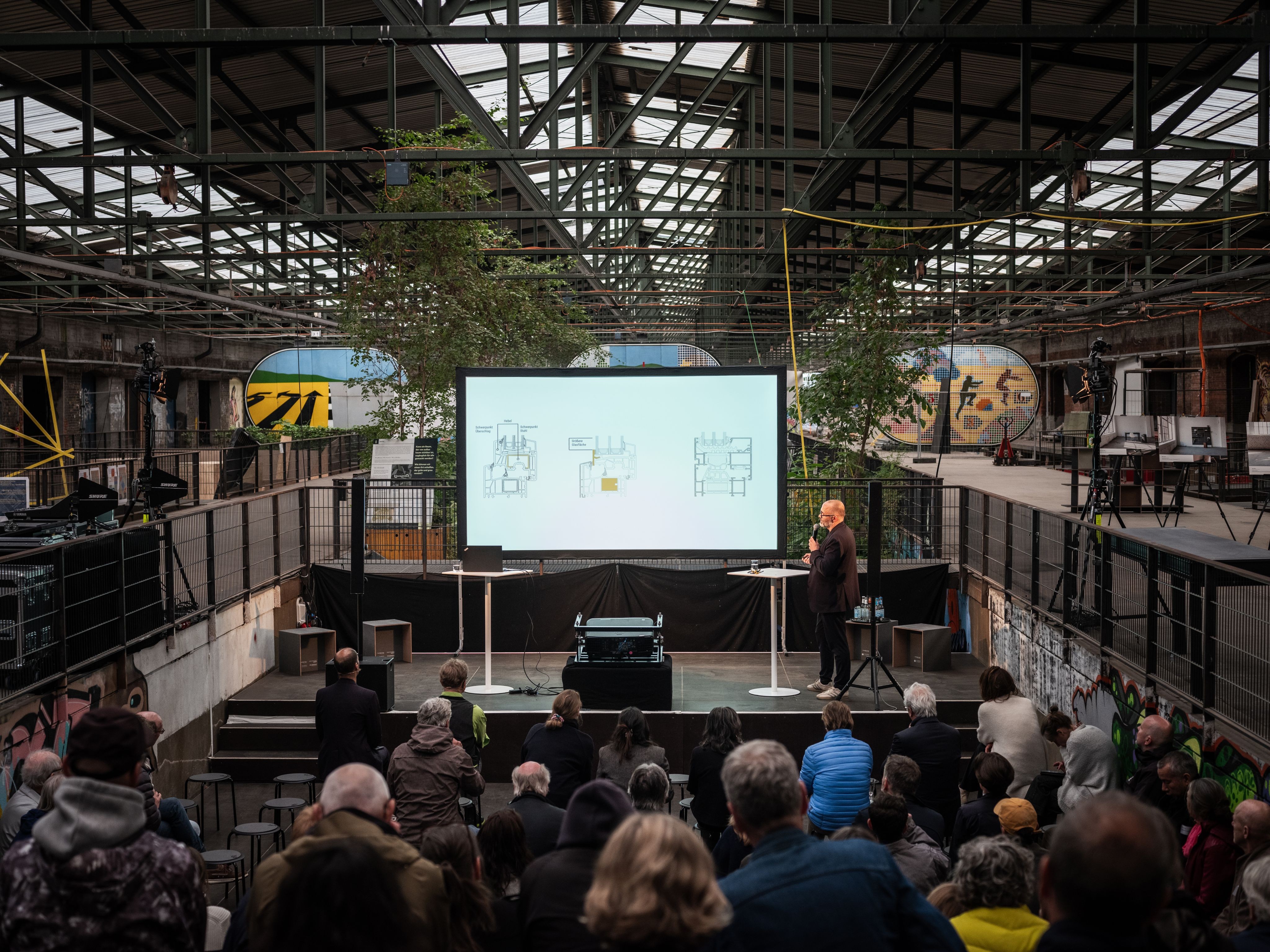 Um homem está em pé no palco, em frente a uma tela grande, fazendo uma apresentação para uma multidão de pessoas.