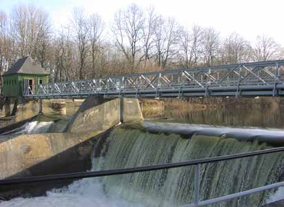 A dam with cascading water and a metal bridge overhead, set against bare trees and a small green building in the background.