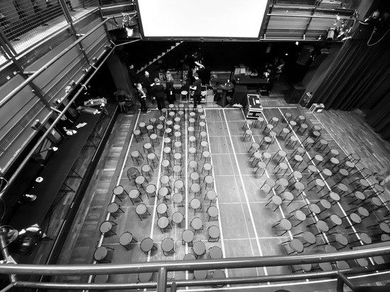 A black and white image of an empty auditorium with chairs arranged in rows, a stage with a large screen, and people standing on the stage.