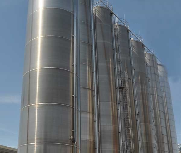 A row of tall, stainless steel silos with ladders and railings against a clear blue sky.