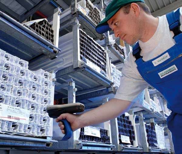 A man in a blue apron and green cap is scanning a barcode on a pallet in a warehouse.