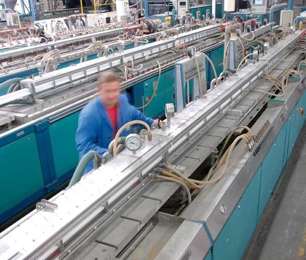A man in a blue jacket is working on a machine in a factory. He is holding a gauge and looking at the machine. The machine has a long metal frame with pipes and wires attached to it. There are other machines and equipment in the background.