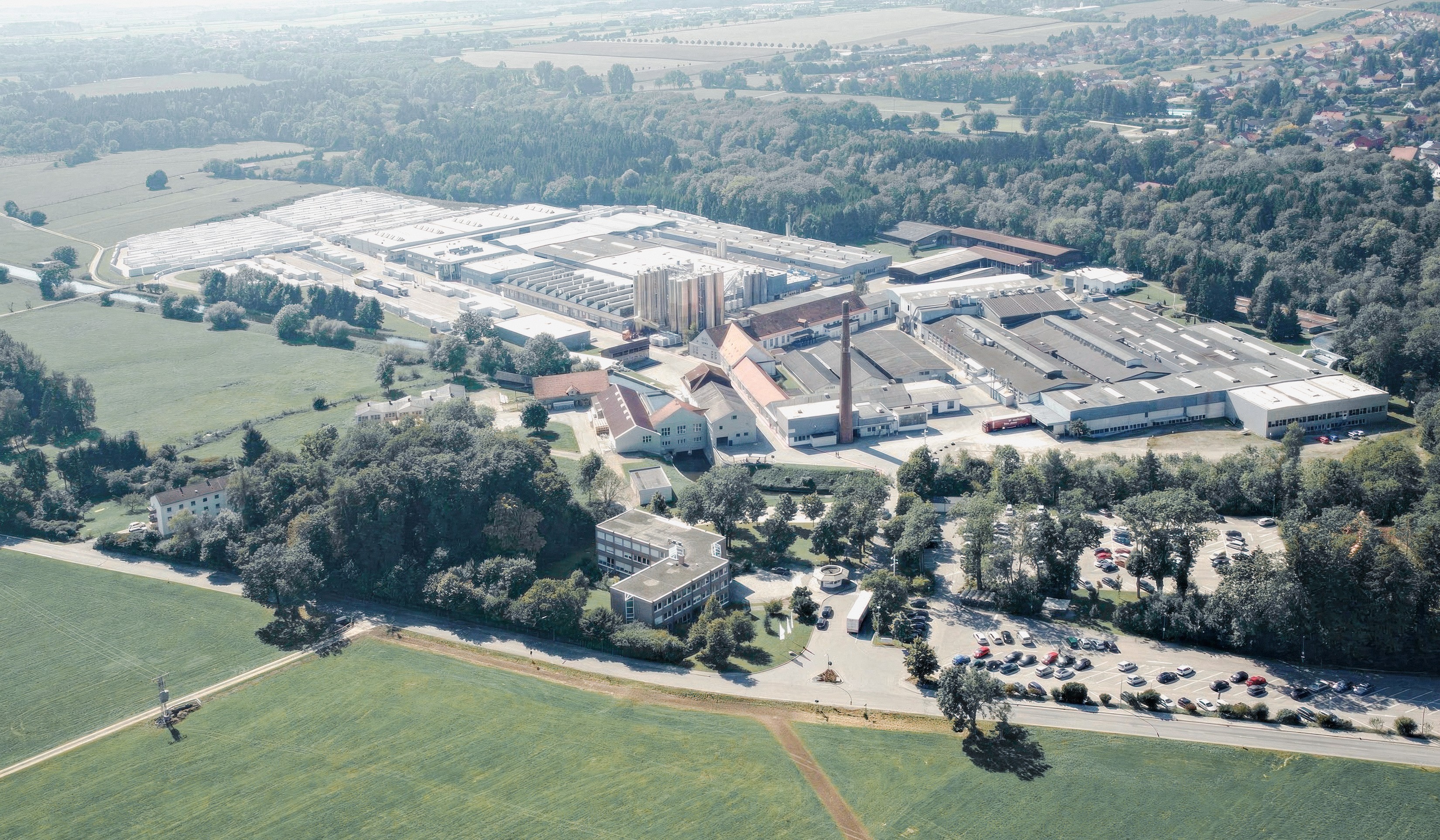 Luchtfoto van een groot industrieel complex met meerdere gebouwen, een hoge schoorsteen en een parkeerplaats vol met auto's. Het complex is omgeven door bomen en velden.