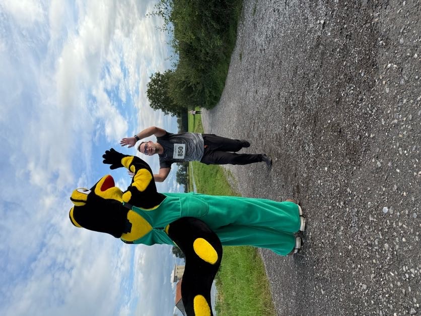 A man wearing a race bib with the number 850 runs down a gravel road with a mascot.