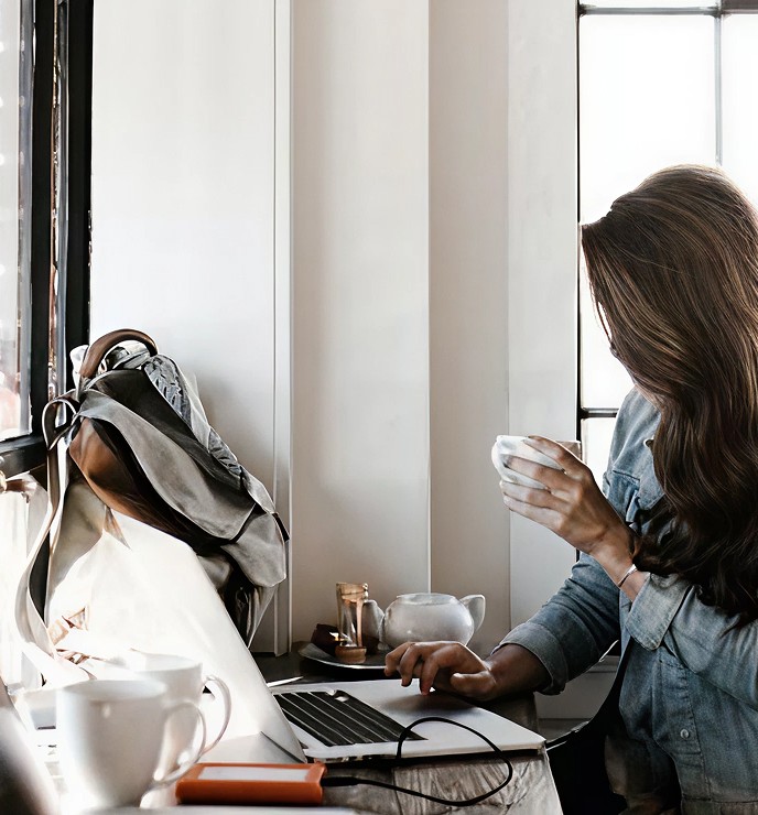Eine Frau sitzt an einem Tisch mit einem Laptop, trinkt aus einer Tasse und schaut aus dem Fenster.