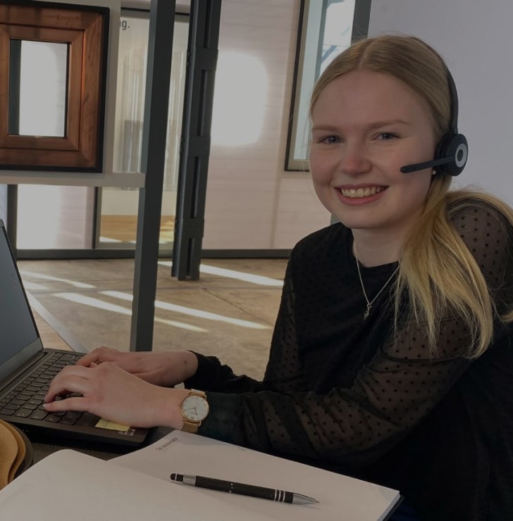 Eine blonde Frau, die ein Headset-Mikrofon und eine schwarze Bluse mit schwarzen Punkten trägt, sitzt vor einem Laptop. Sie lächelt und tippt auf der Laptoptastatur. Ein Stift und ein Stück Papier liegen auf dem Tisch vor ihr. Hinter ihr befindet sich eine Glaswand mit einem Holzrahmen und eine weiße Wand.