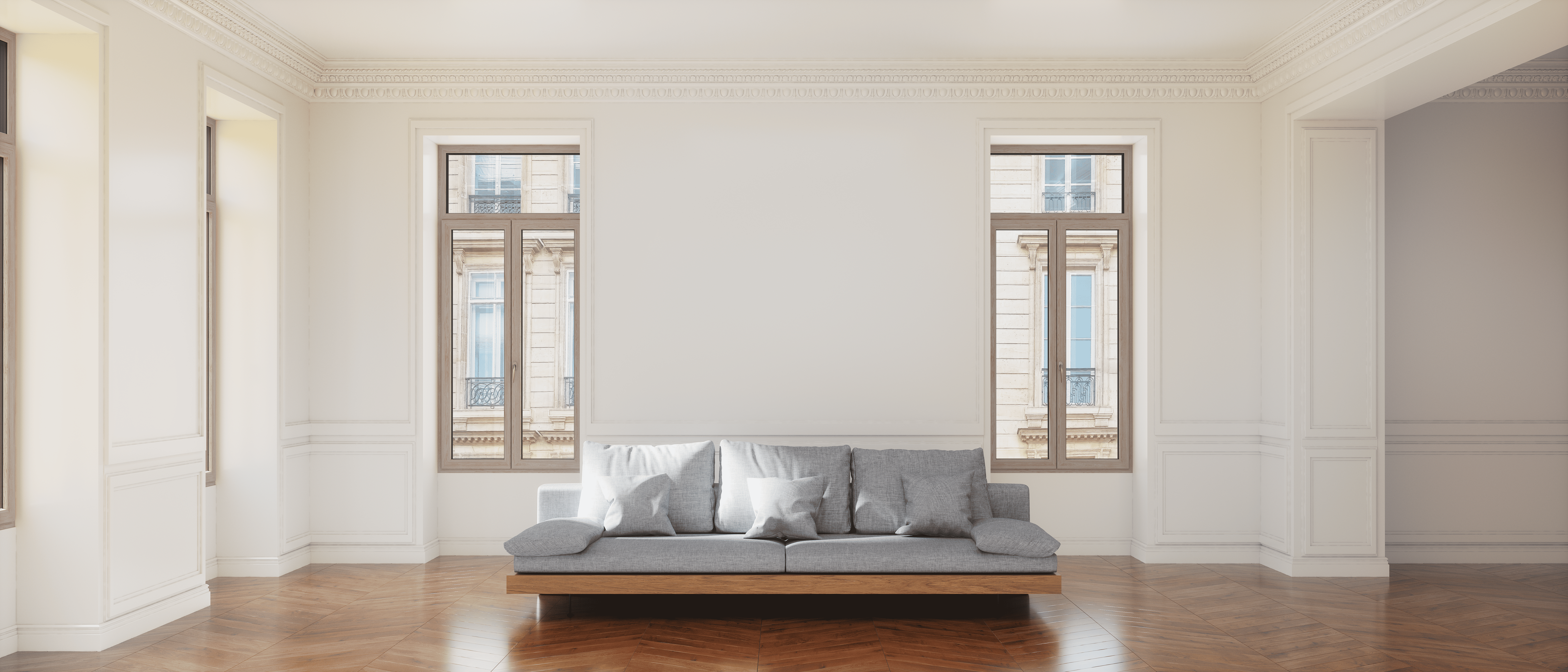 Un divano grigio con quattro cuscini è posizionato di fronte a due grandi finestre in una stanza con pareti bianche e pavimenti in legno.