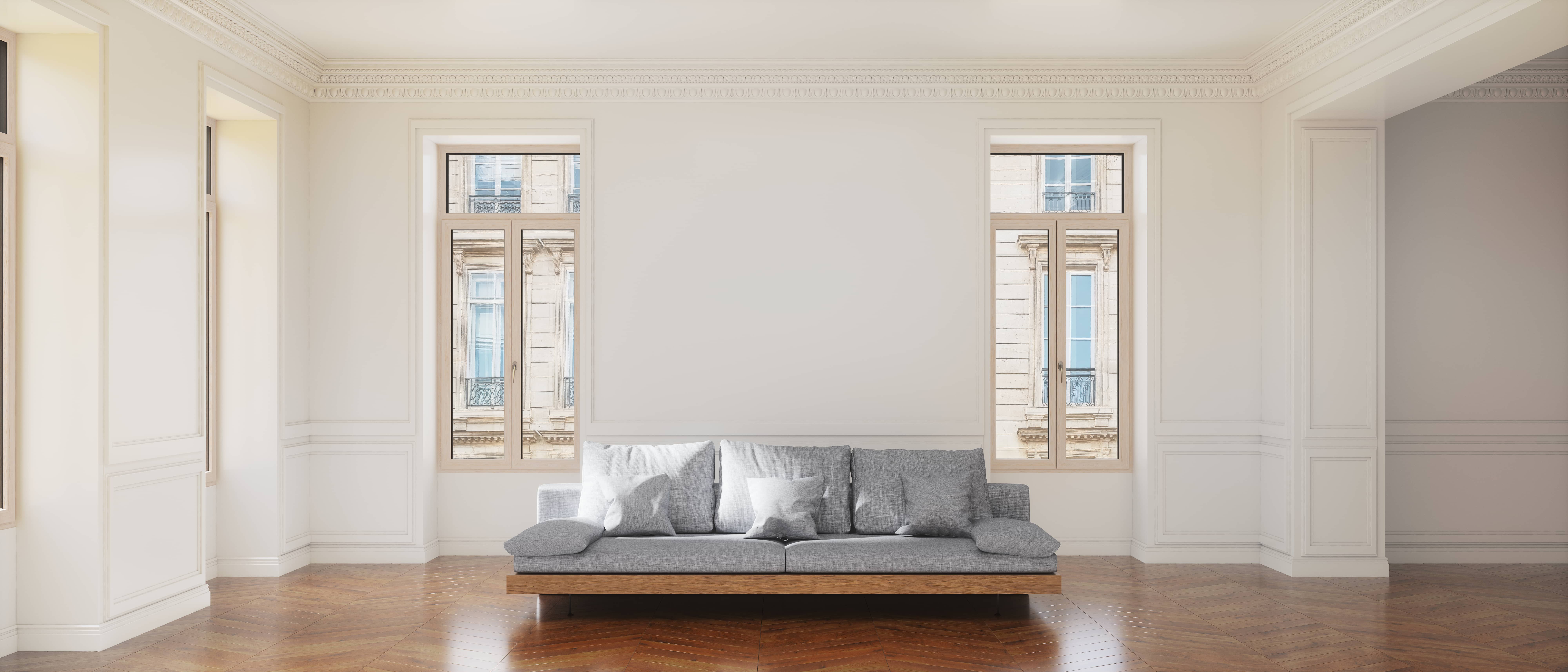 Een grijze sofa met vier kussens staat in het midden van een kamer met witte muren, houten vloeren en twee grote ramen.