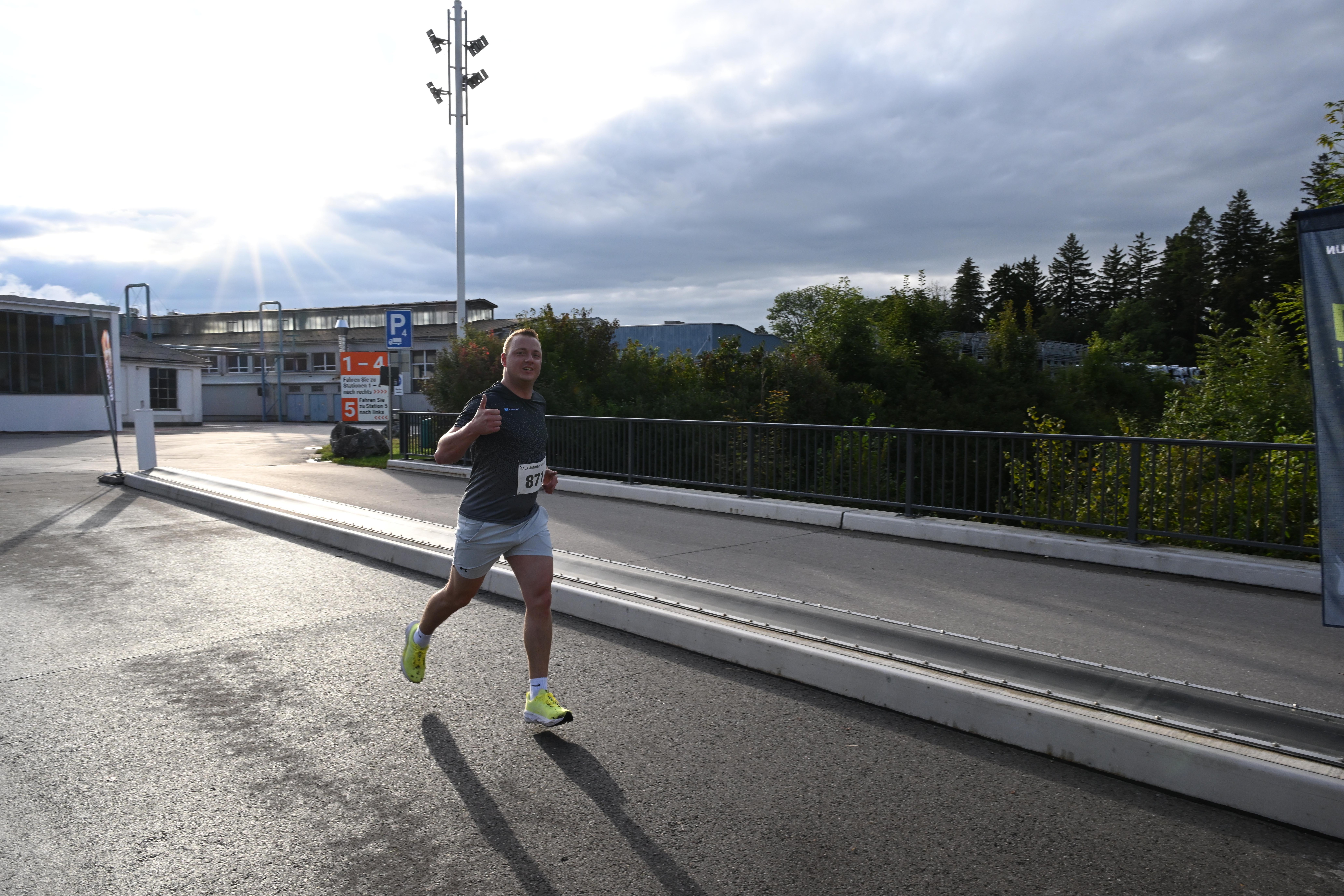 A man running on a road with a thumbs up, wearing a gray t-shirt, white shorts, and yellow sneakers. Behind him, there is a metal fence, a parking sign, and a building. In the distance, there are trees and a cloudy sky.