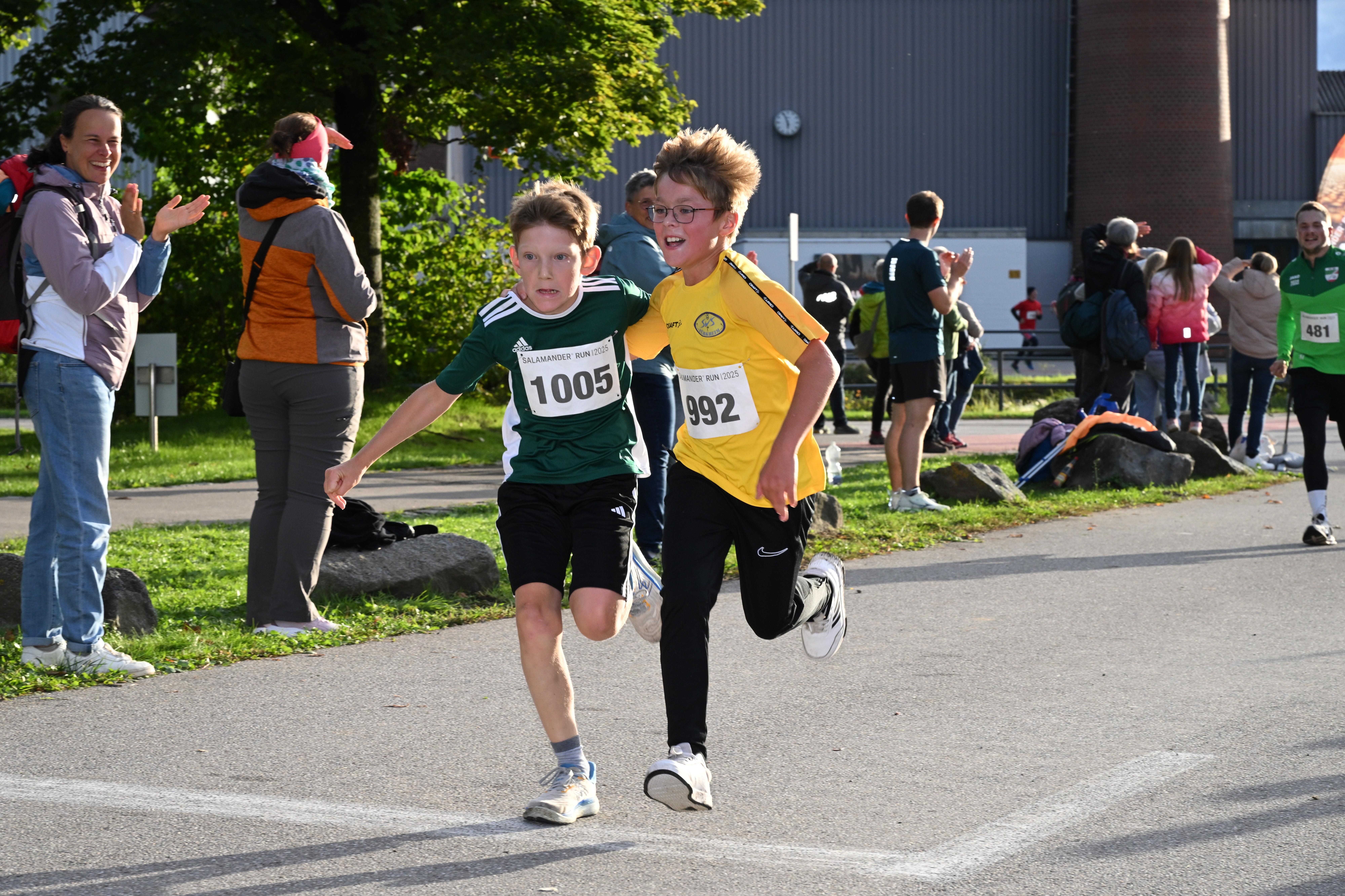 Two boys running in a race with numbers pinned to their shirts. One boy wears a green shirt and the other a yellow shirt. Spectators are cheering them on from the sidelines.