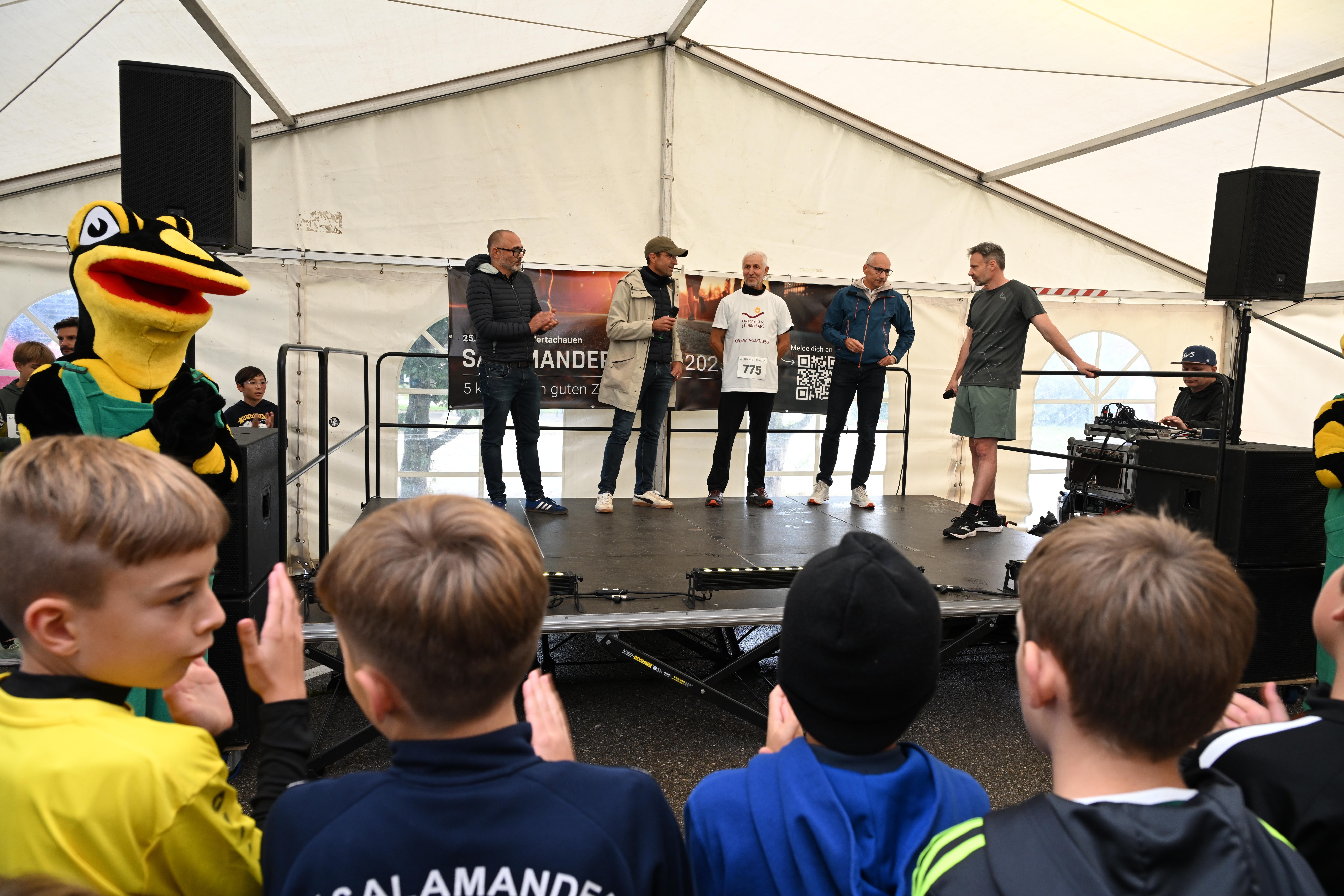 A group of men standing on a stage in front of a crowd of children. The men are wearing casual clothing and some are holding a banner. The children are clapping and cheering. The stage is set up with speakers and a mascot. The event appears to be a celebration or announcement.