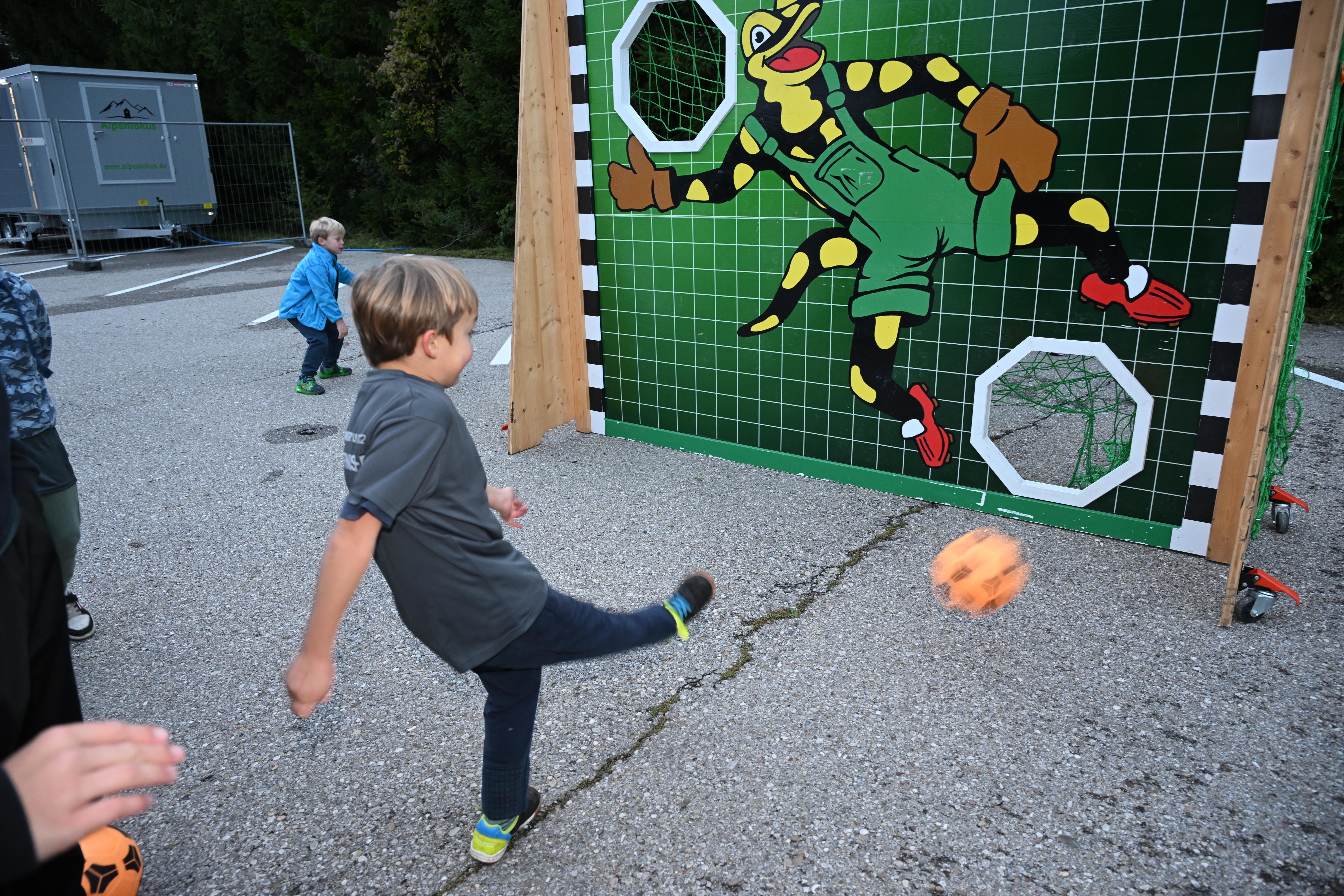 A boy kicks a ball towards a goal with a frog goalie on it while other children play in the background.