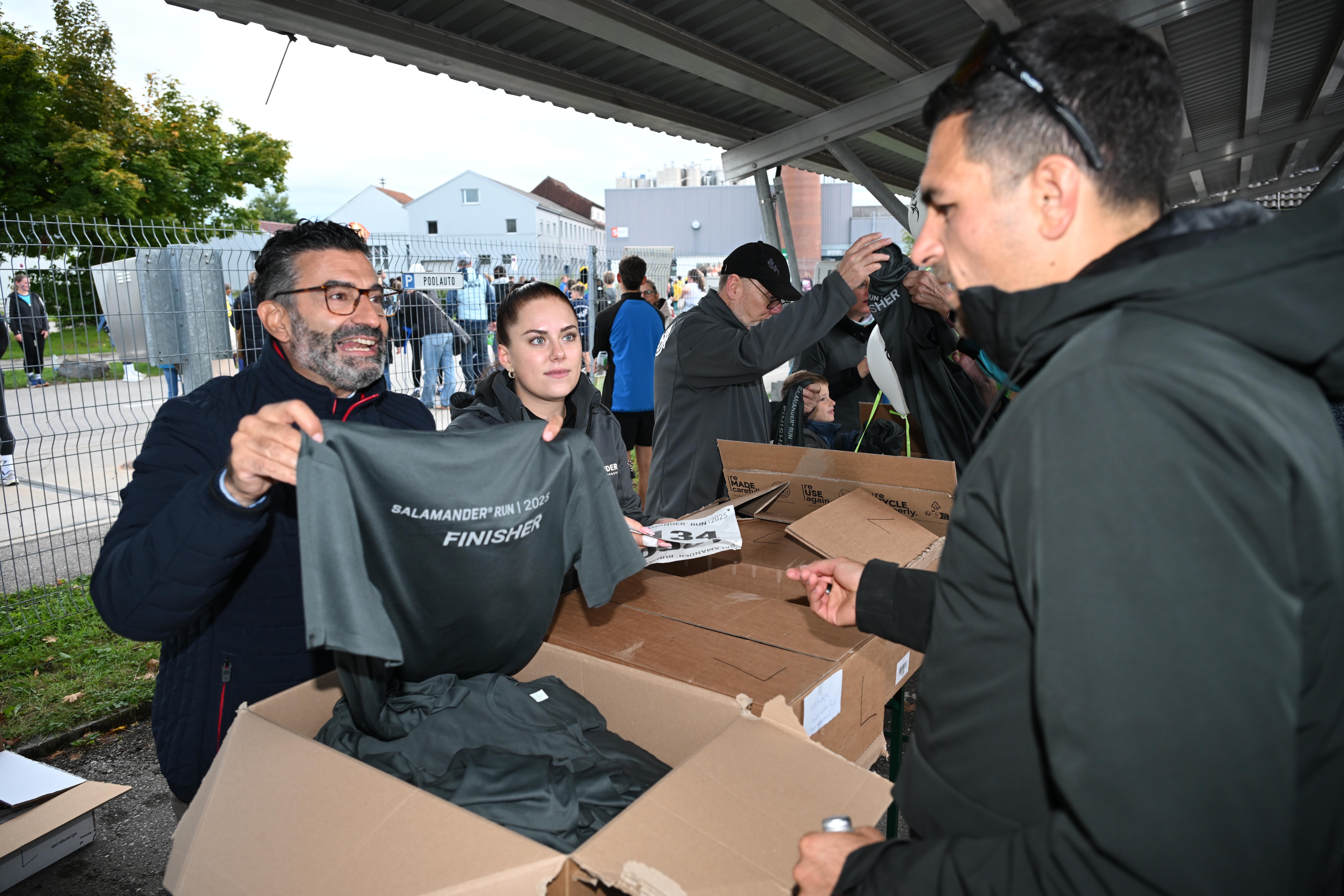 A man and a woman are standing next to a box of merchandise. The man is holding a shirt and the woman is holding a box. They are both wearing jackets and glasses. Behind them, there are other people standing and sitting on the ground. There is a fence and a building in the background.