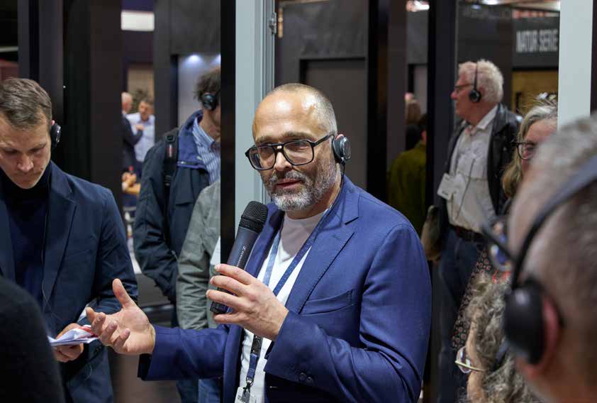 A man wearing glasses speaks into a microphone at an event, surrounded by people wearing headsets.