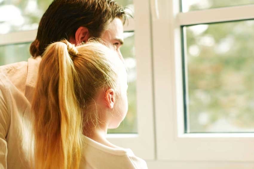 A man and a young girl with a ponytail are standing close together and looking out of a window.