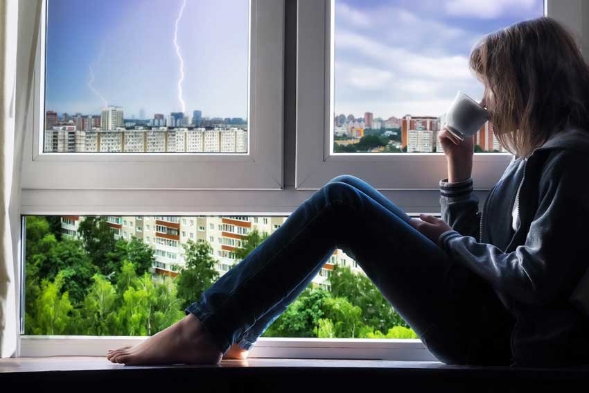 A woman is sitting on a windowsill, drinking from a mug, and watching a lightning storm through the window.