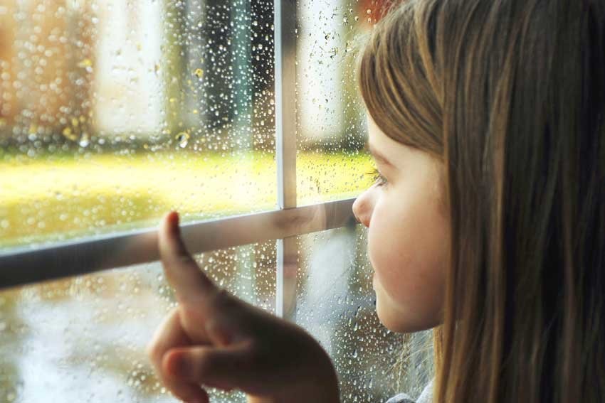 A young girl with long hair is looking out of a window during a rainy day.