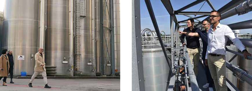 People walking near large steel tanks; others standing on a metal platform, observing industrial equipment under a clear sky.