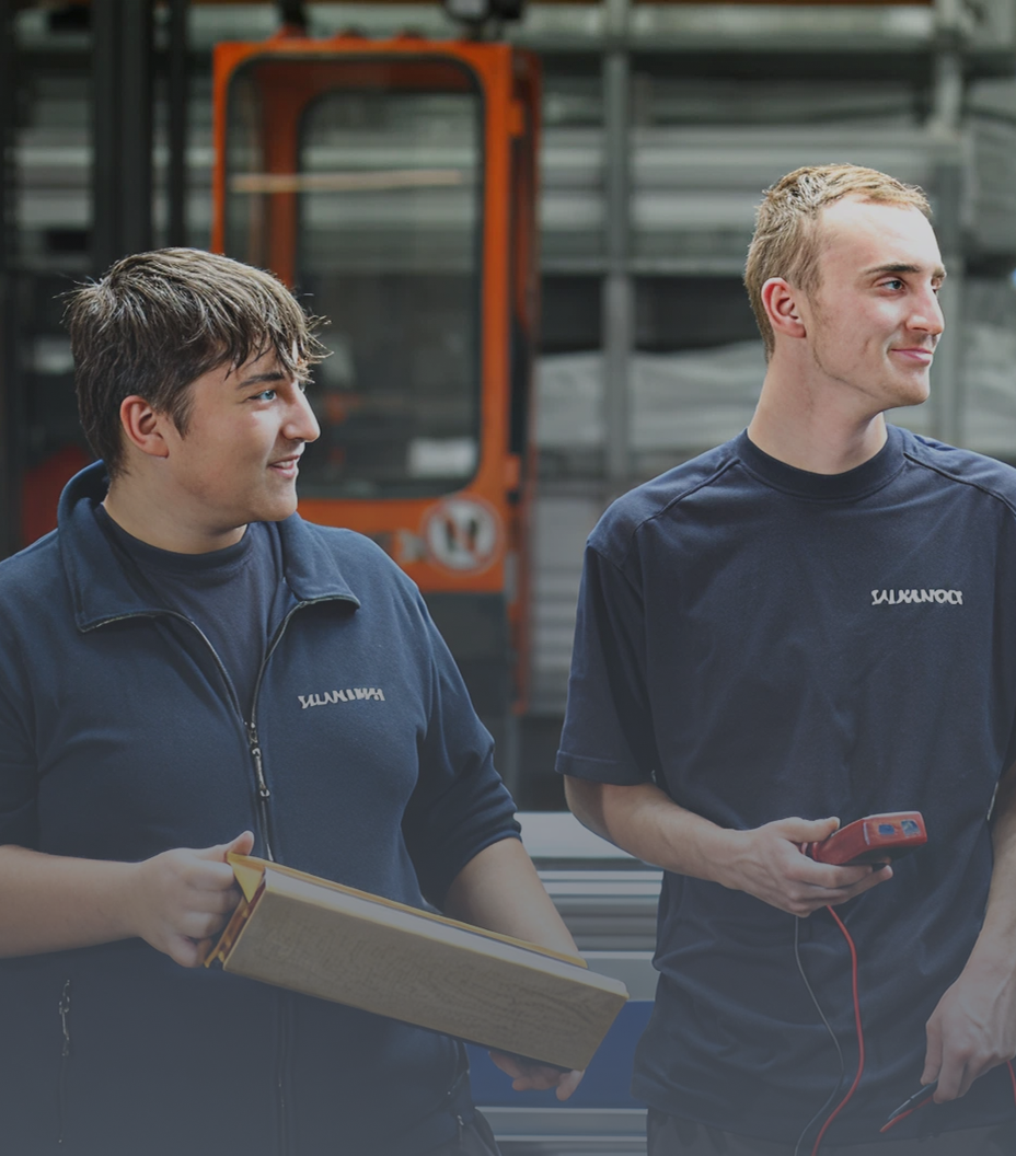 Zwei Männer in blauen Uniformen stehen in einer Fabrik, einer hält eine Kiste und der andere ein rotes Gerät.