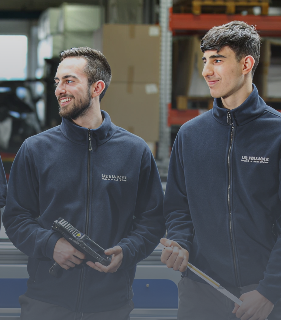 Zwei Männer in blauen Jacken stehen nebeneinander in einem Lagerhaus. Einer hält ein Werkzeug, und der andere hält ein Maßband.