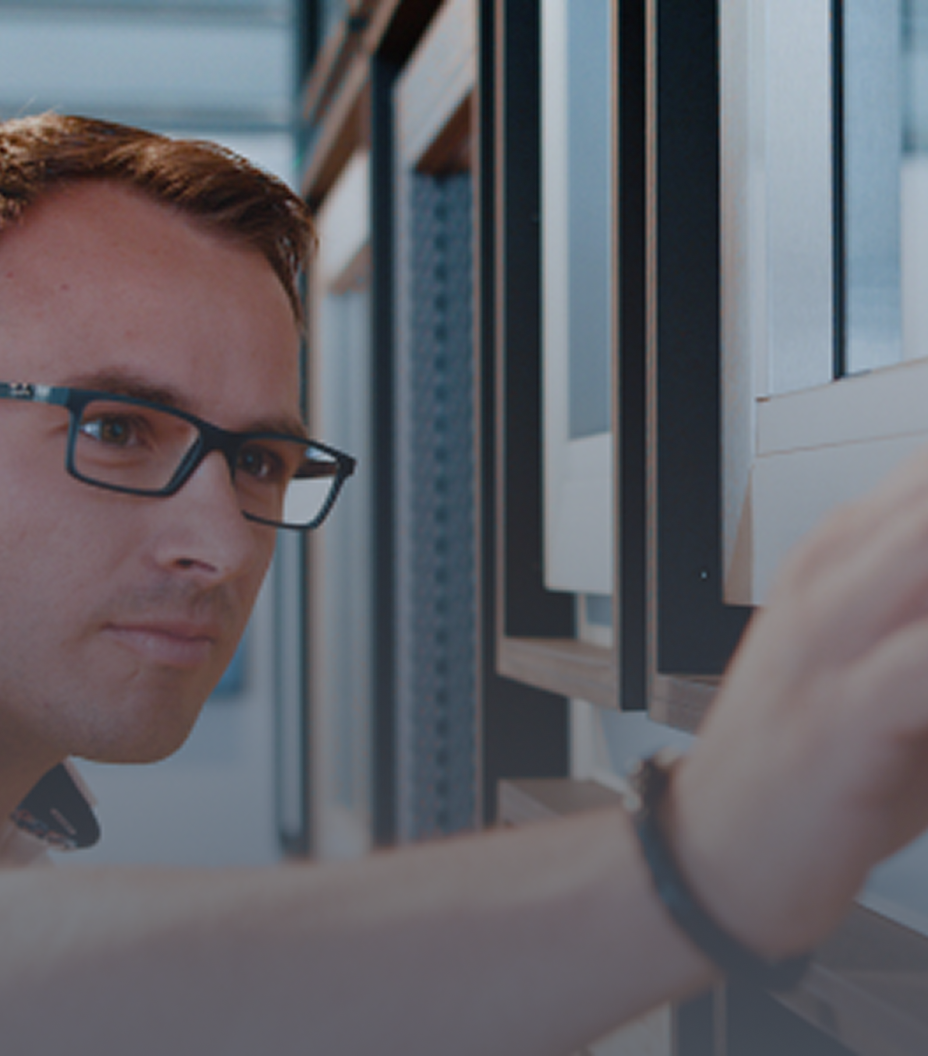 Ein Mann mit Brille und Armband steht vor einer Glaswand und berührt sie mit seiner rechten Hand.