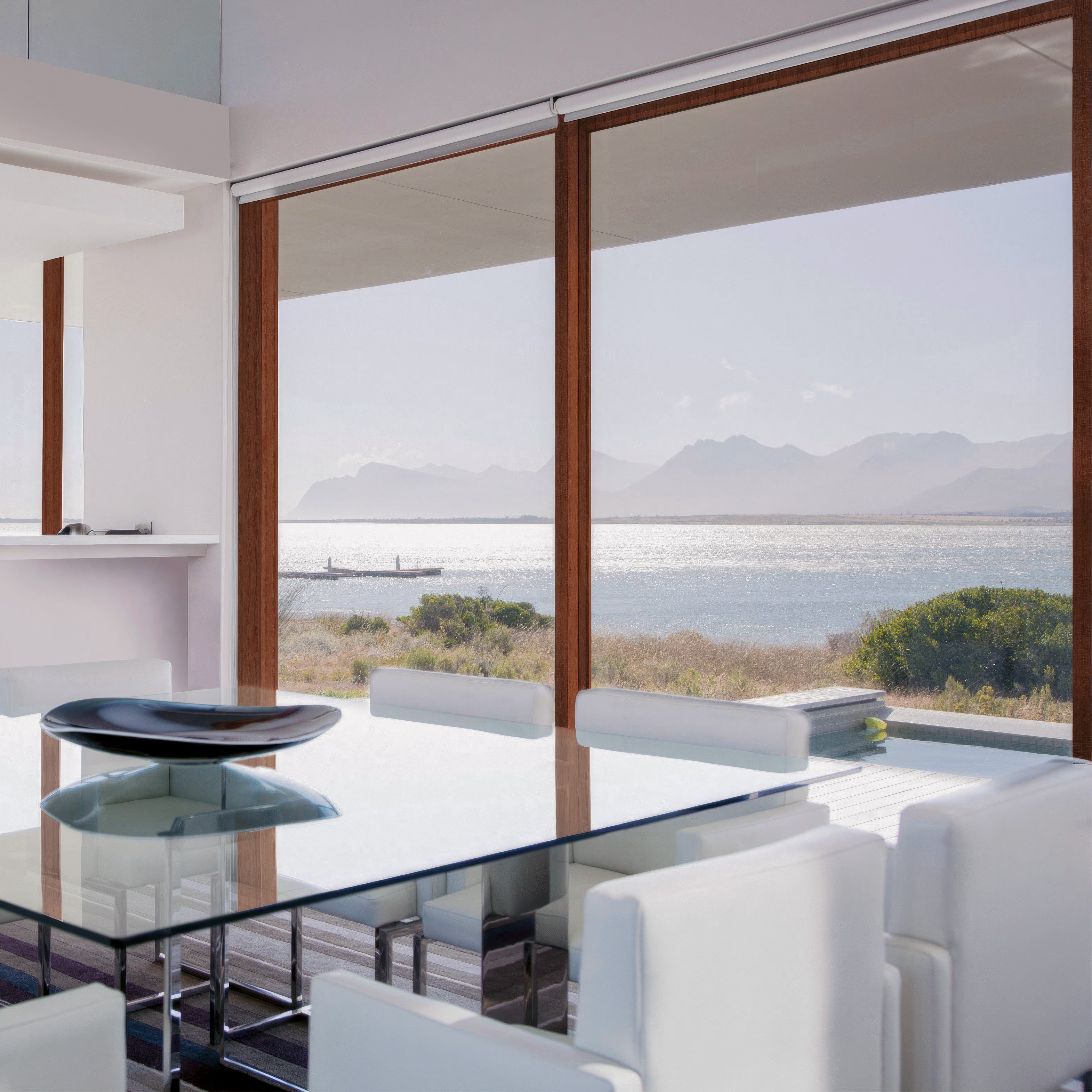 Un comedor con una mesa de vidrio y sillas blancas, con vista a una piscina y al océano.