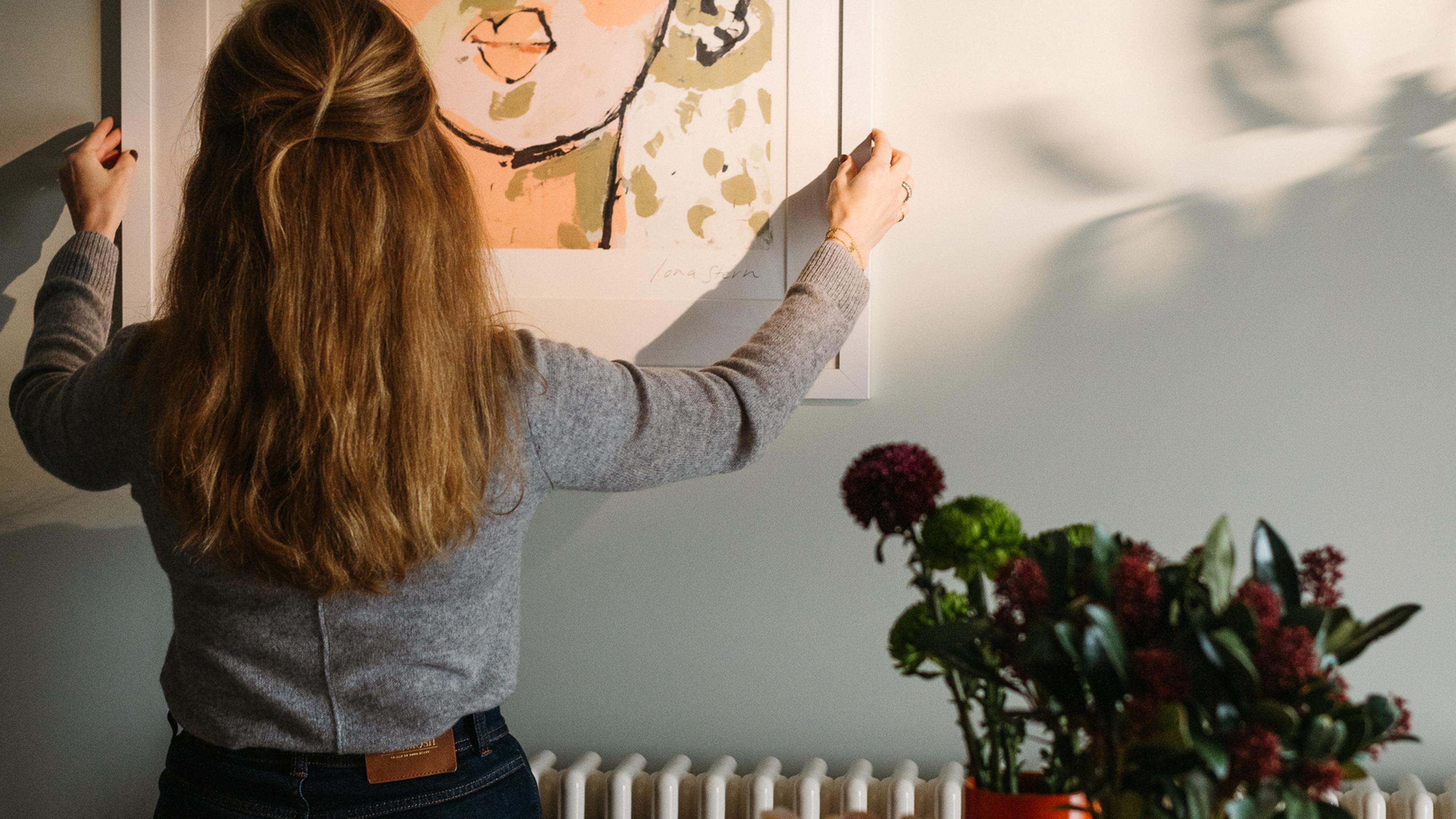 A woman is hanging a print of a face of a man in dappled sunlight.