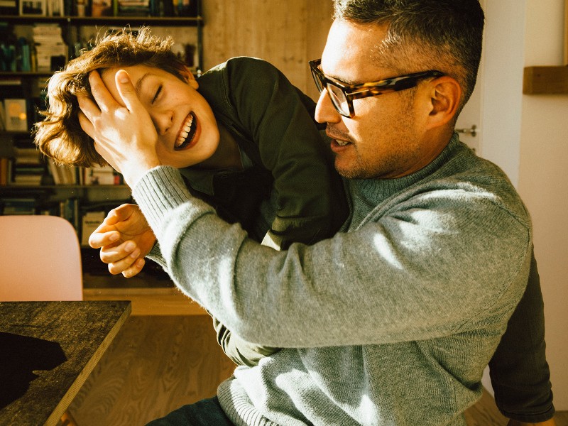 Vater mit Sohn. Der Vater stützt sein Gesicht mit der Hand, während sie zusammen spielen. Beide lachen fröhlich.
