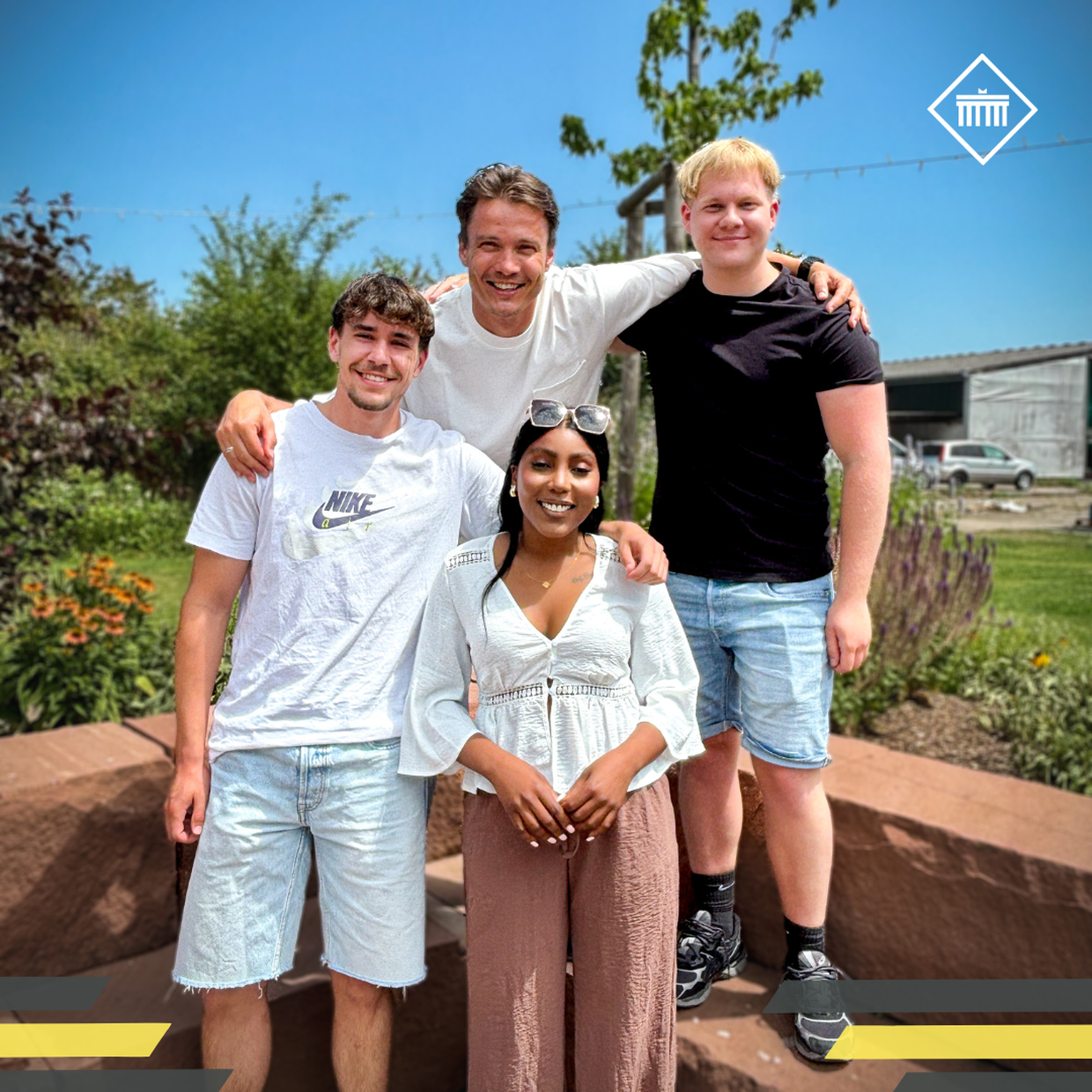 Four people smiling outdoors, with greenery and a building in the background. Three men and one woman, all casually dressed.