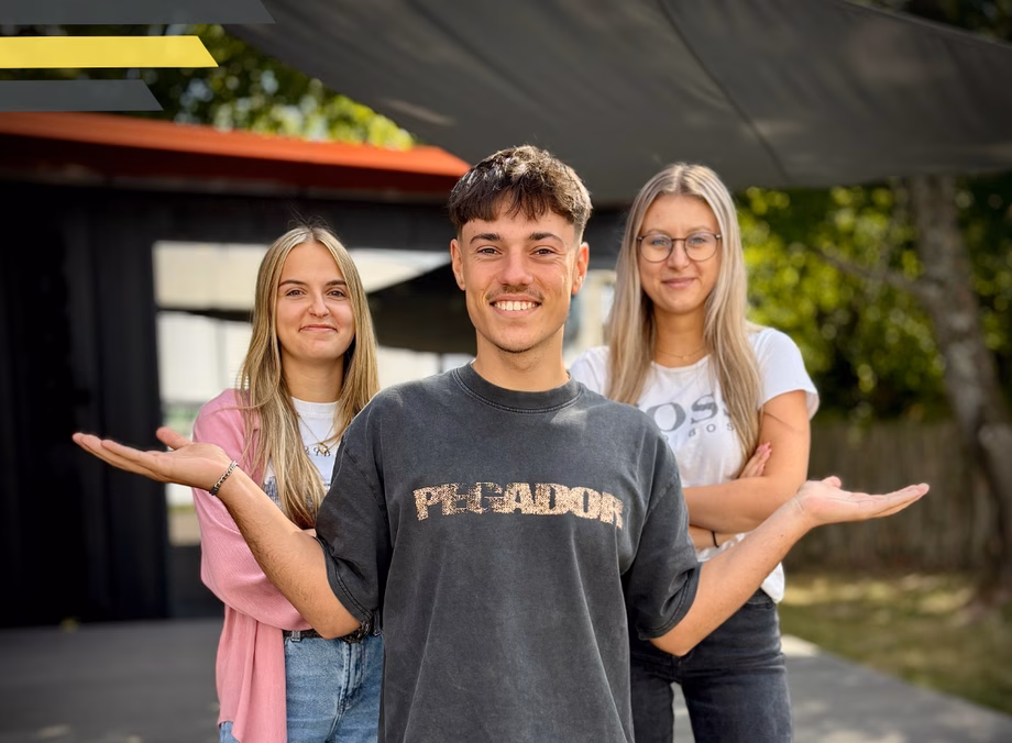 Three smiling people stand outdoors, with one person in front gesturing with open hands. A building and trees are in the background.
