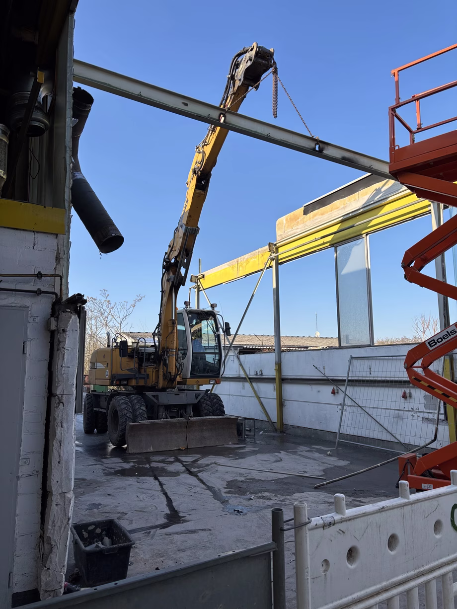 A construction site with a yellow excavator lifting a beam, surrounded by scaffolding and barriers under a clear blue sky.