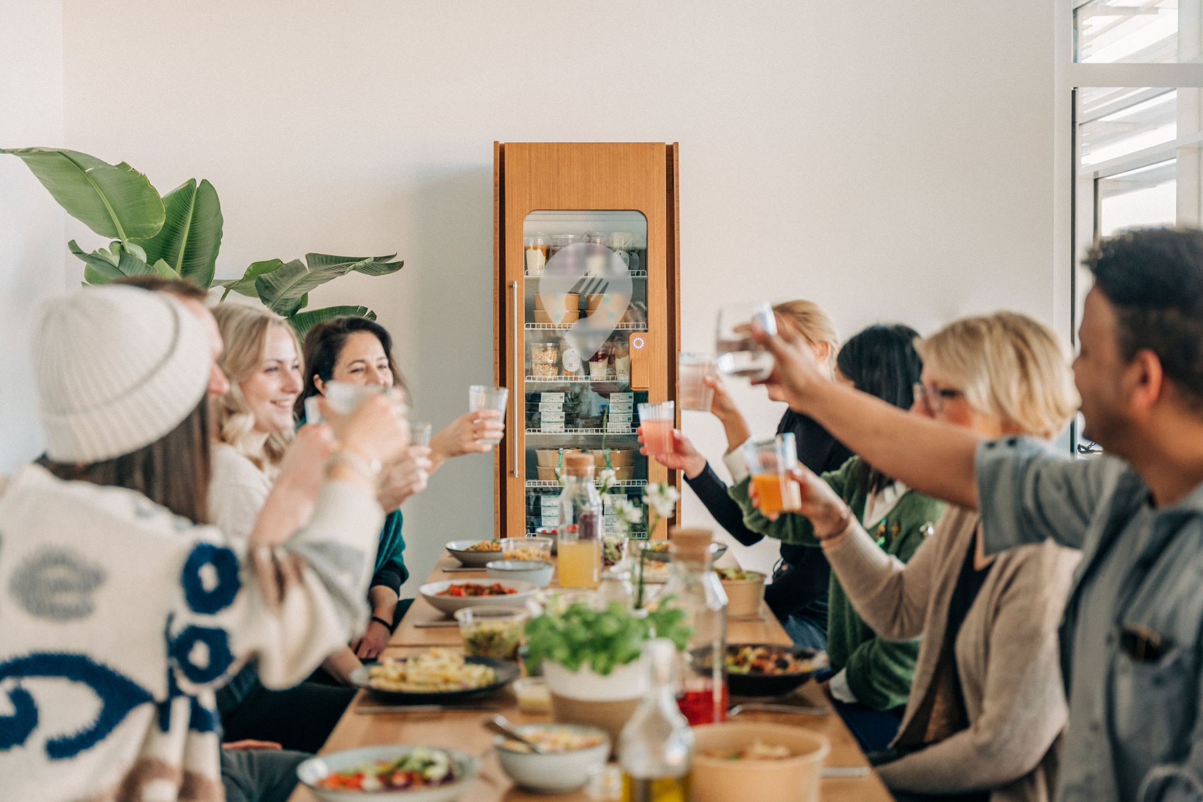 A group of people sit around a table, toasting with drinks, surrounded by plates of food in a bright room with plants and a cabinet. A group of people sit around a table, toasting with drinks, surrounded by plates of food in a bright room with plants and a cabinet.